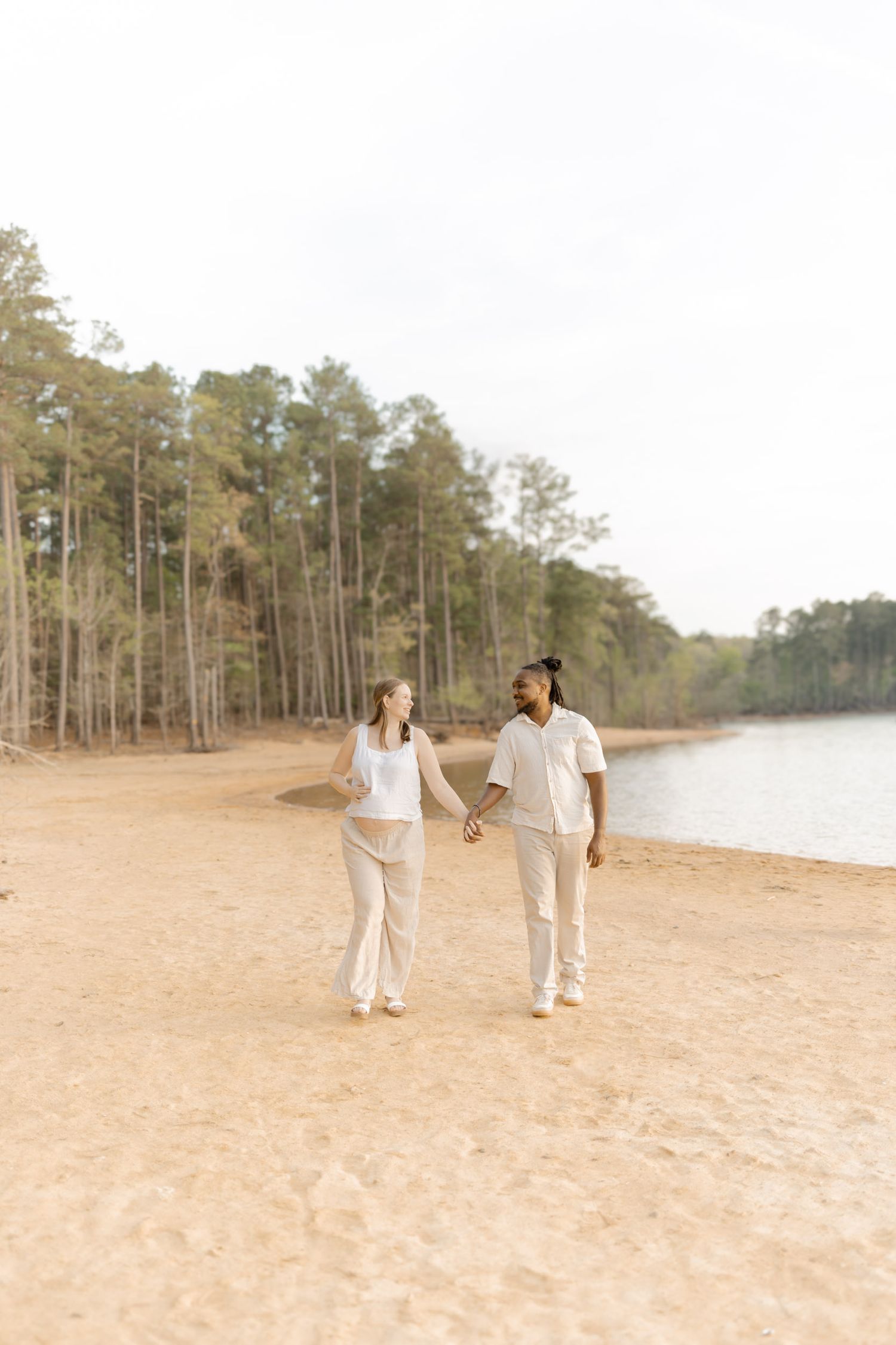 Couple holding hands walking on sandy beach shore with tall pine trees and calm water in background.