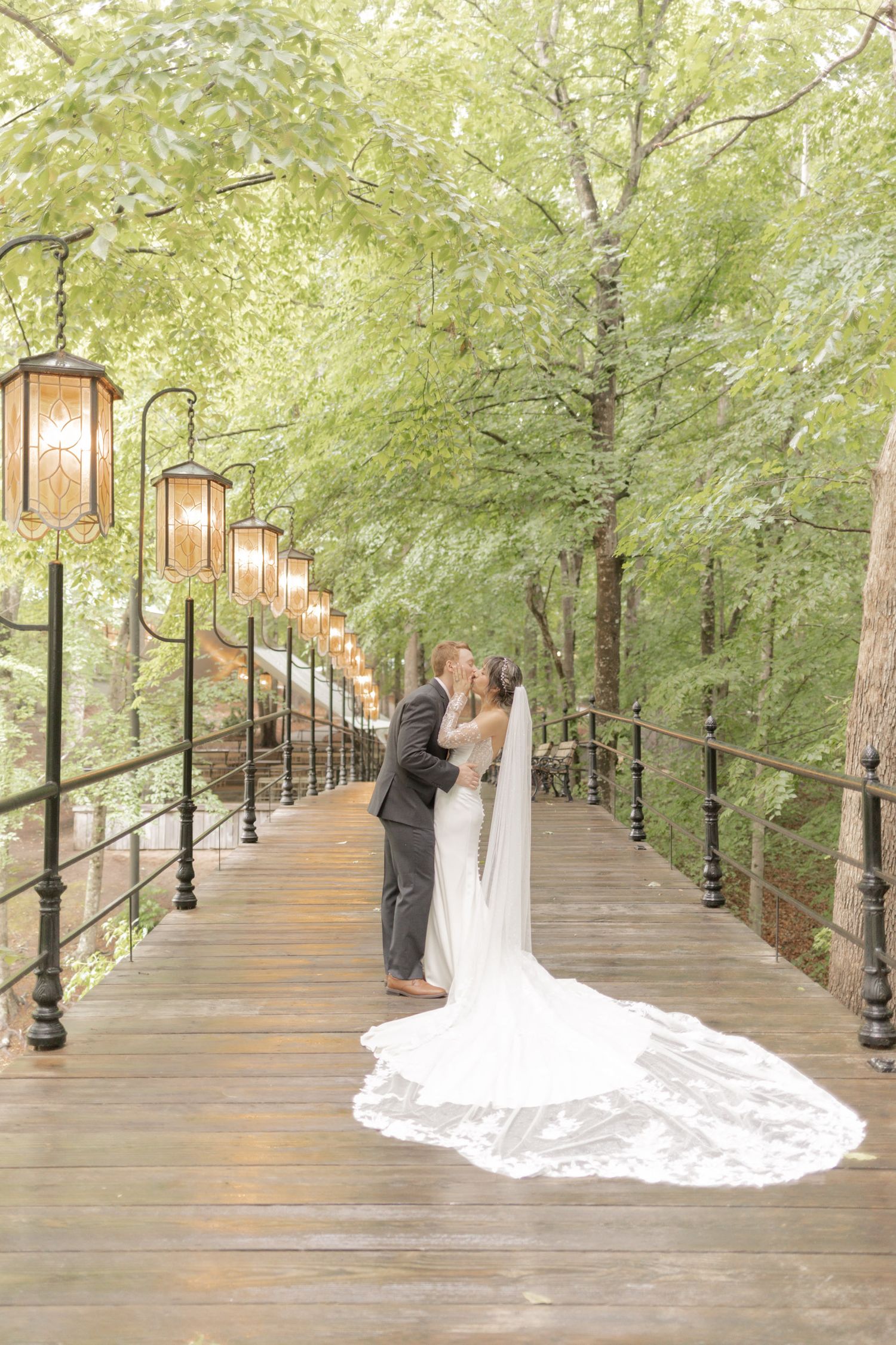 Bride and groom kissing on a lantern-lit wooden bridge surrounded by lush green trees, bride's veil trailing elegantly.