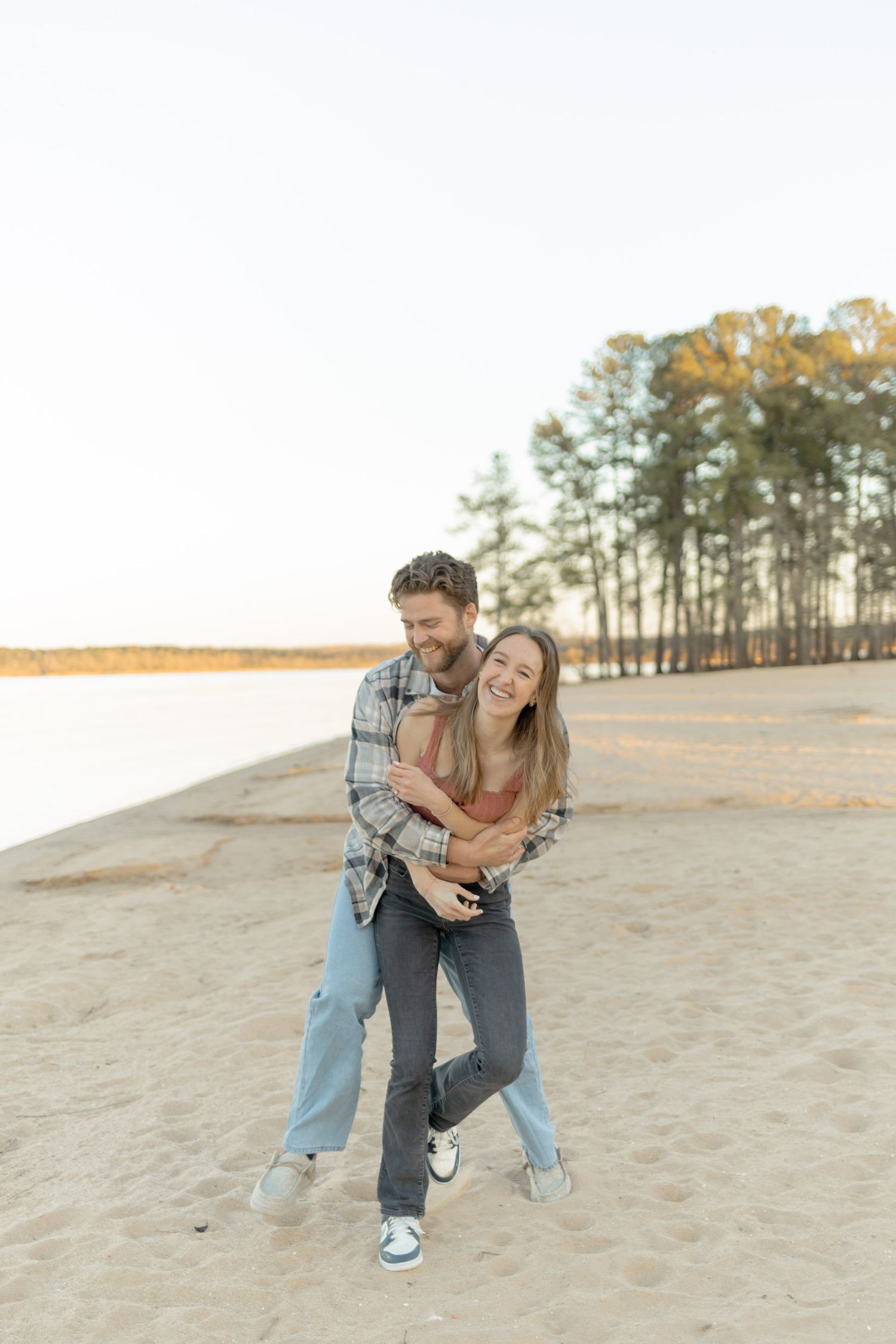 A couple embracing and laughing together on a sandy beach with pine trees in the background during their couples lifestyle photoshoot.
