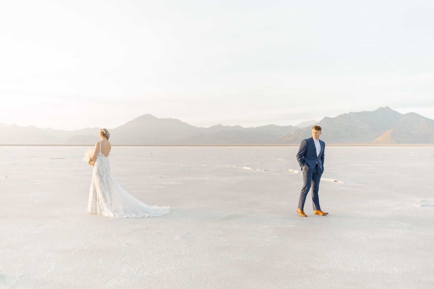 A couple in wedding attire stands apart on a vast salt flat with mountains in the distance during golden hour.