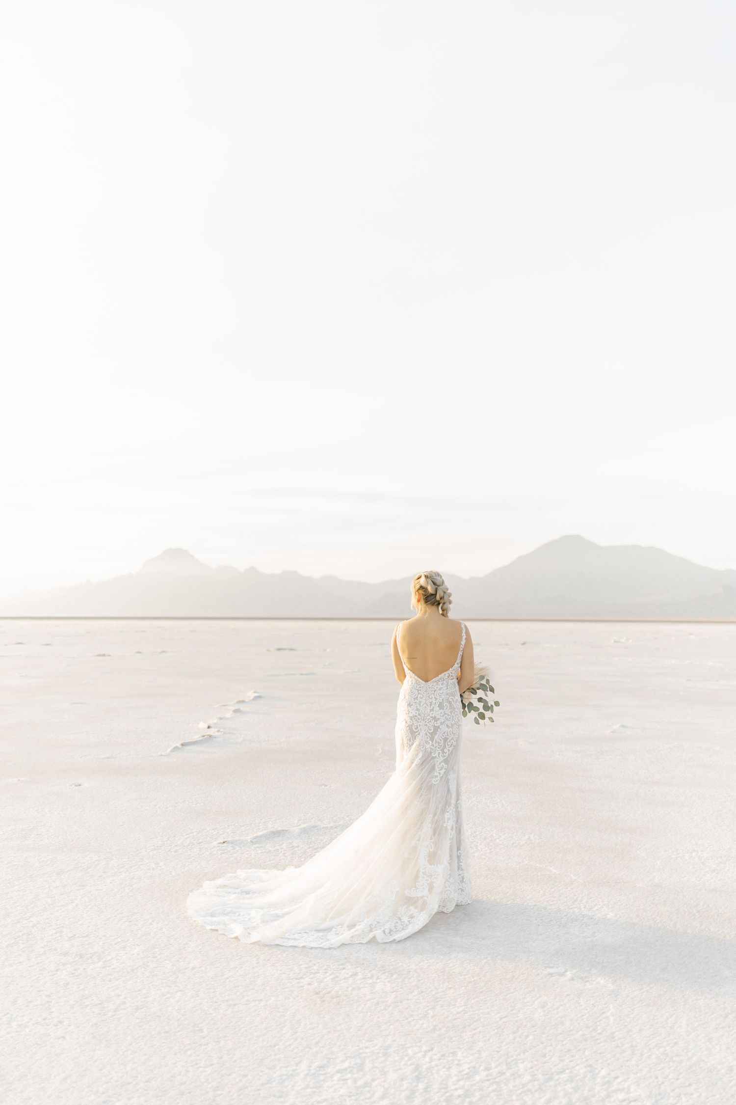 A bride in a flowing white gown walks gracefully across pristine white sand dunes at sunset.