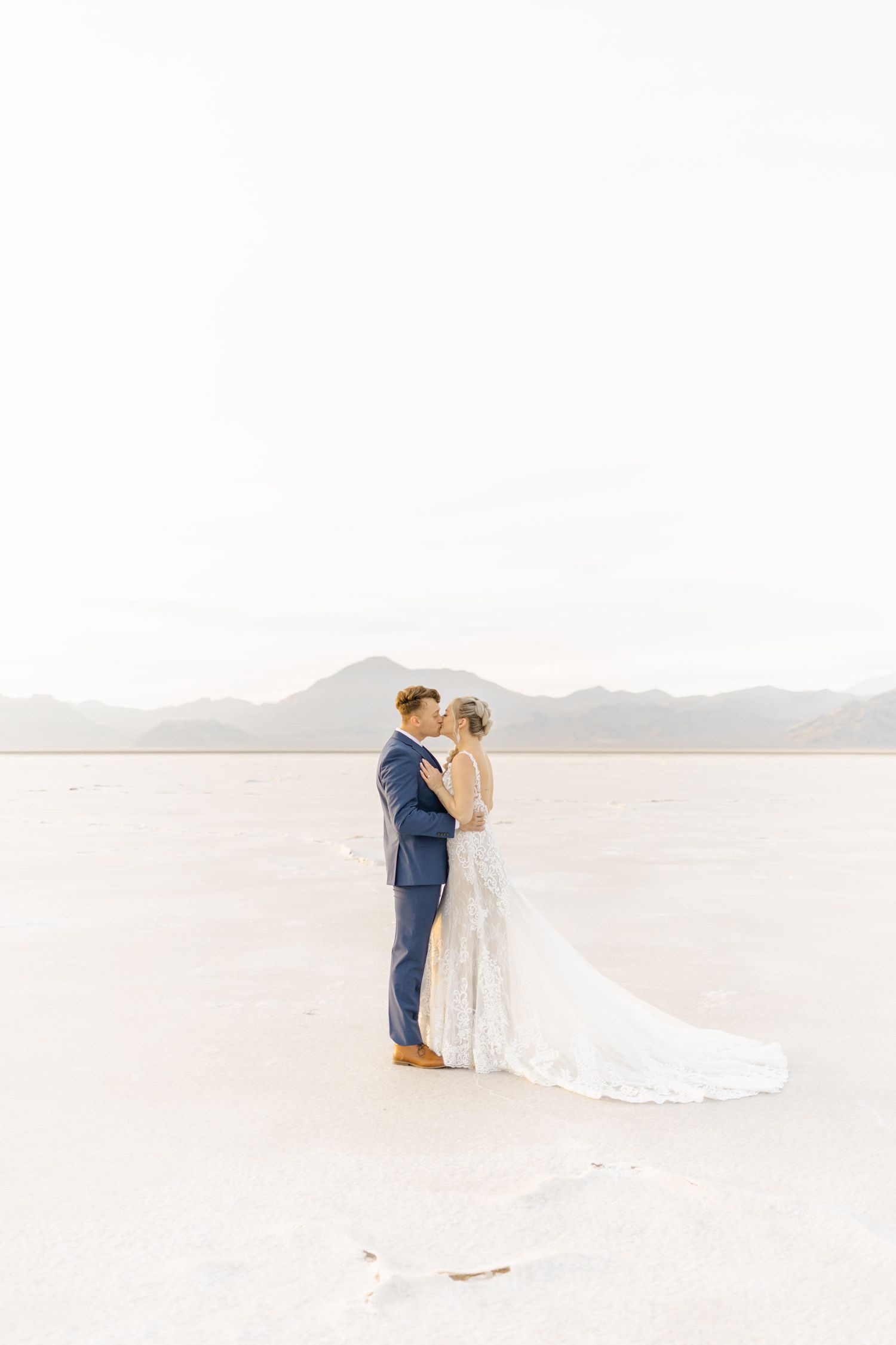 Newlyweds embrace on a stark white salt flat with distant mountains creating a minimalist wedding scene.