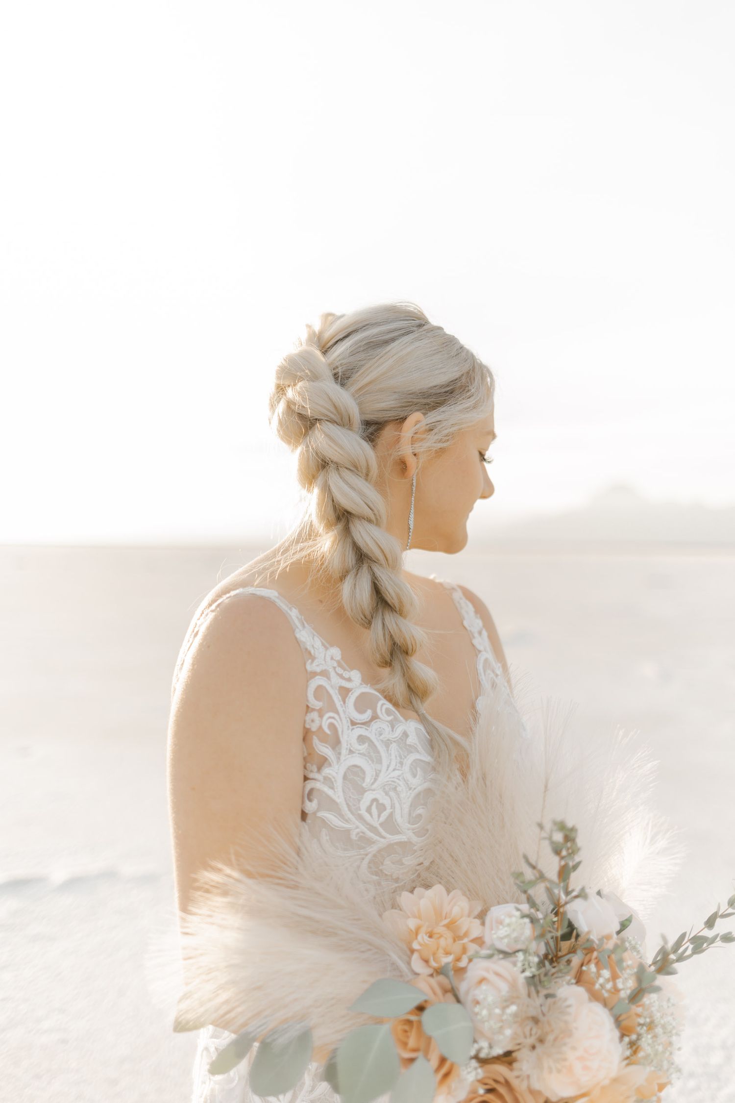 A bride in a lace wedding dress holds a peach and white bouquet against a bright desert landscape.
