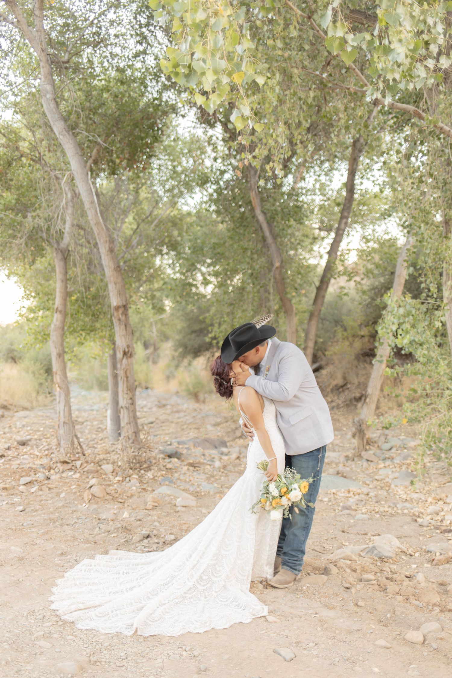 A couple shares a romantic kiss outdoors, bride in lace gown with train, groom in gray shirt and cowboy hat, holding bouquet.