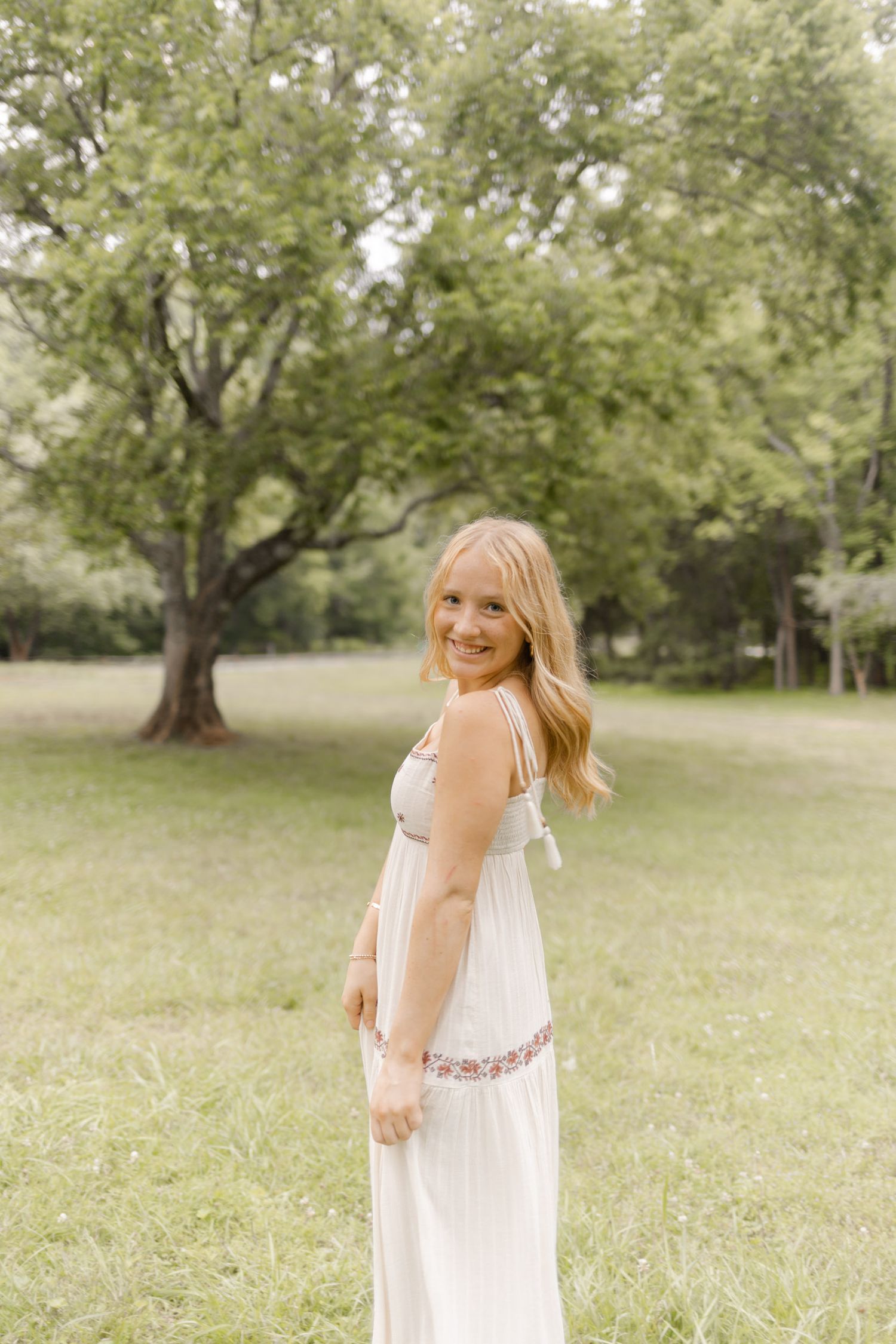 Person in a white summer dress twirls playfully in a sunlit meadow surrounded by trees in Durham, North Carolina.