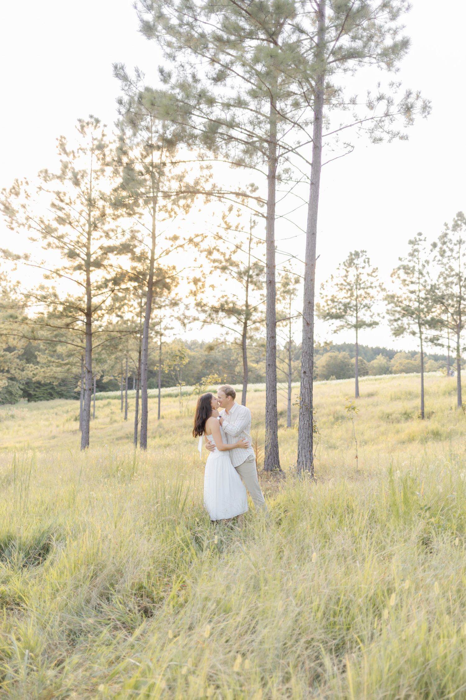 Couple dancing together in a dreamy meadow with pine trees at golden hour during engagement session with photographer in North Carolina.