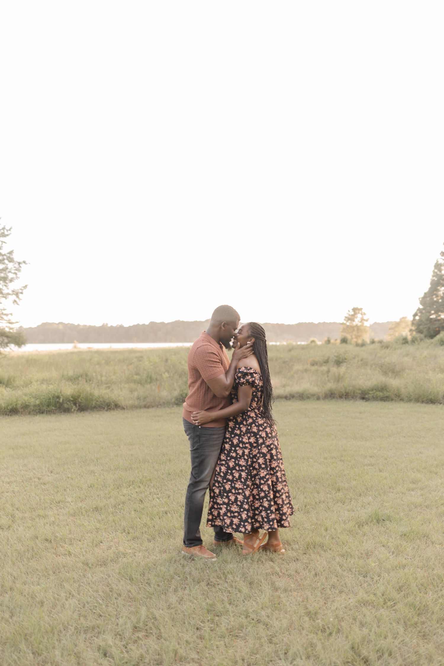 A romantic couple shares a tender embrace while standing in an open grassy field at golden hour.