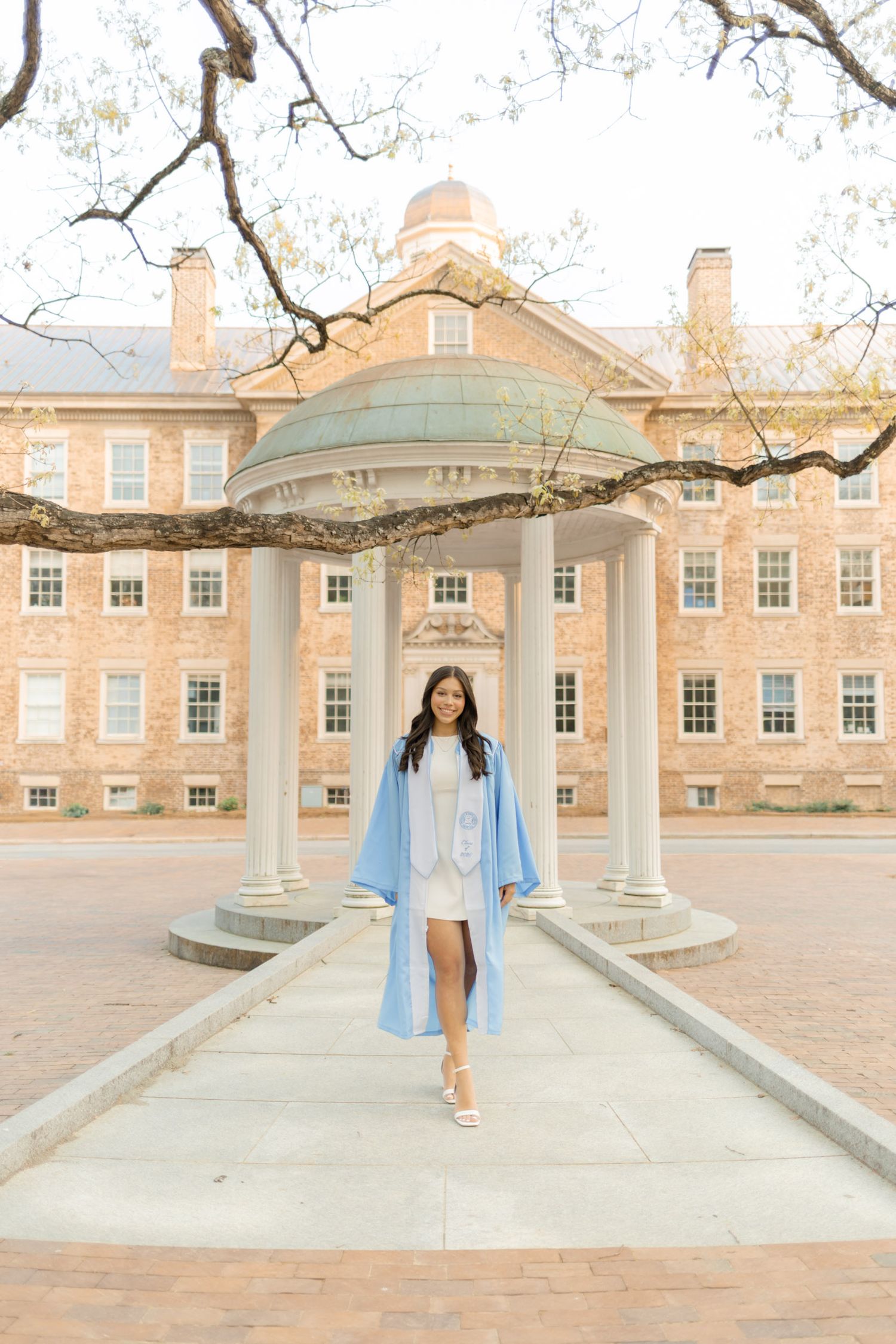 Graduate in light blue cap and gown posing at UNC Chapel Hill Old Well landmark.