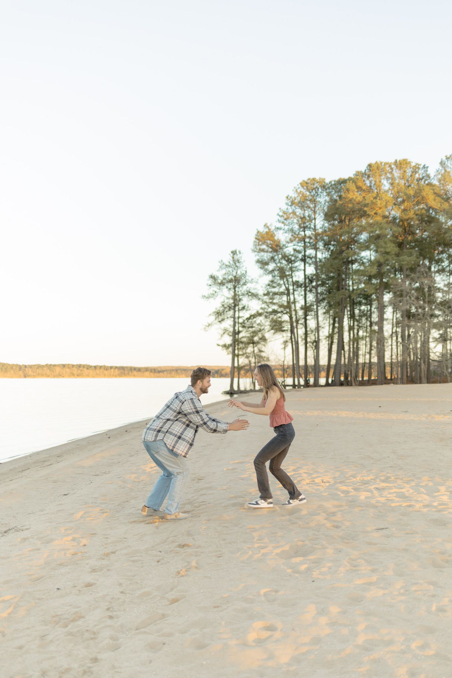 Playful couple running and jumping on sandy dunes with pine trees in background during their couples lifestyle photoshoot in Raleigh.