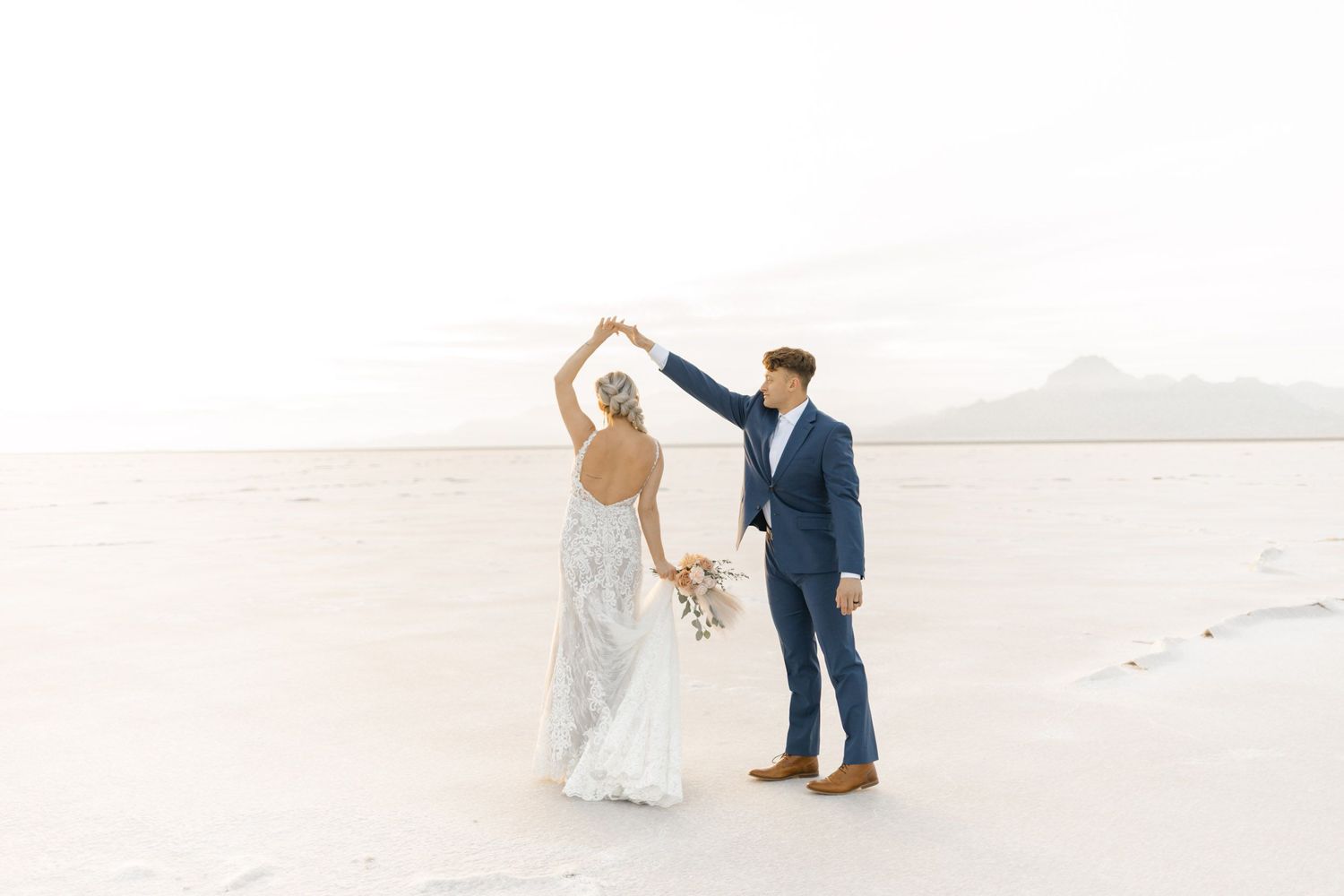 A couple dances together on a bright white salt flat with mountains in the background.
