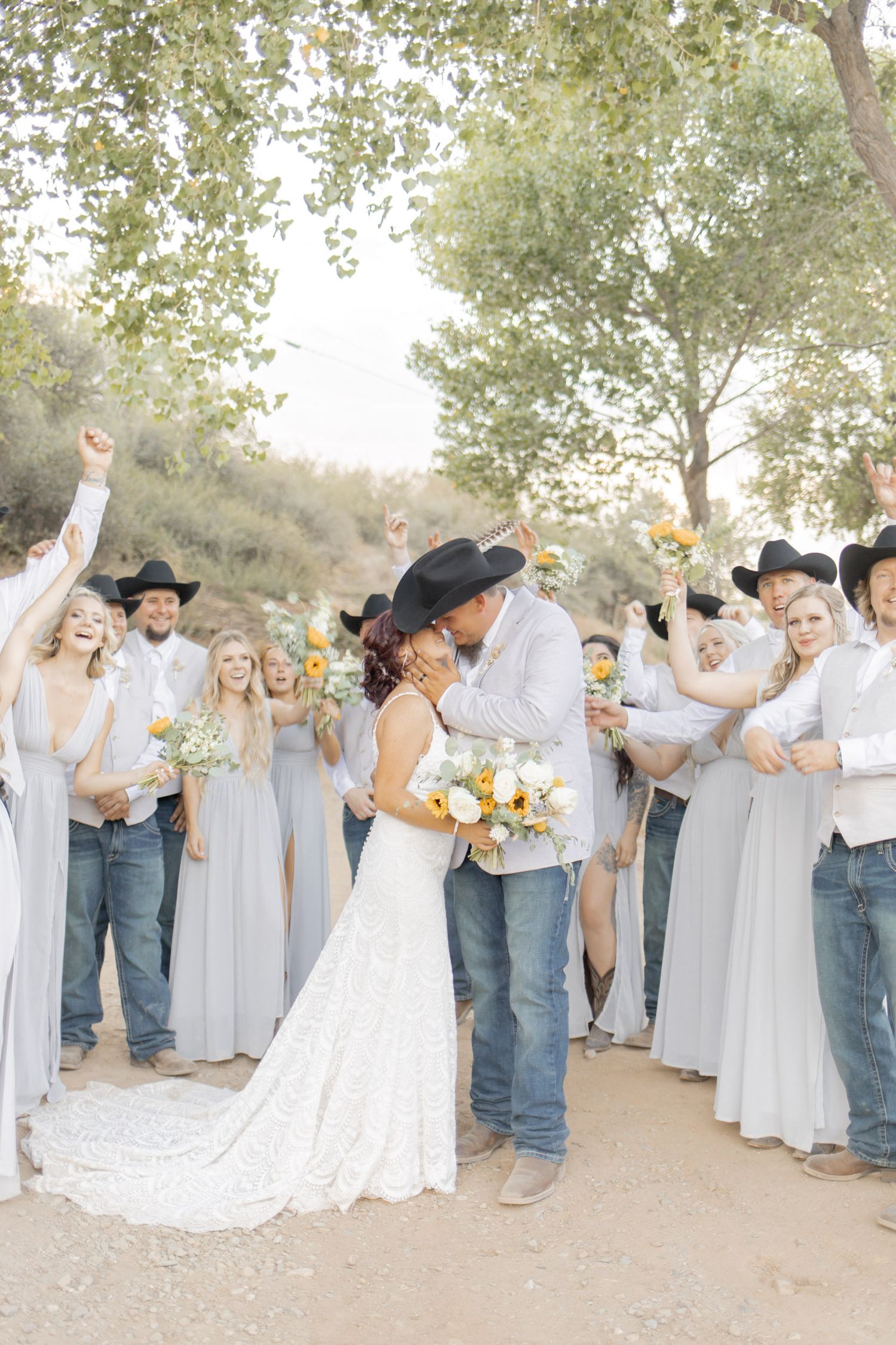 Couple shares a kiss surrounded by cheering wedding party in cowboy hats, jeans, and gray dresses outdoors.