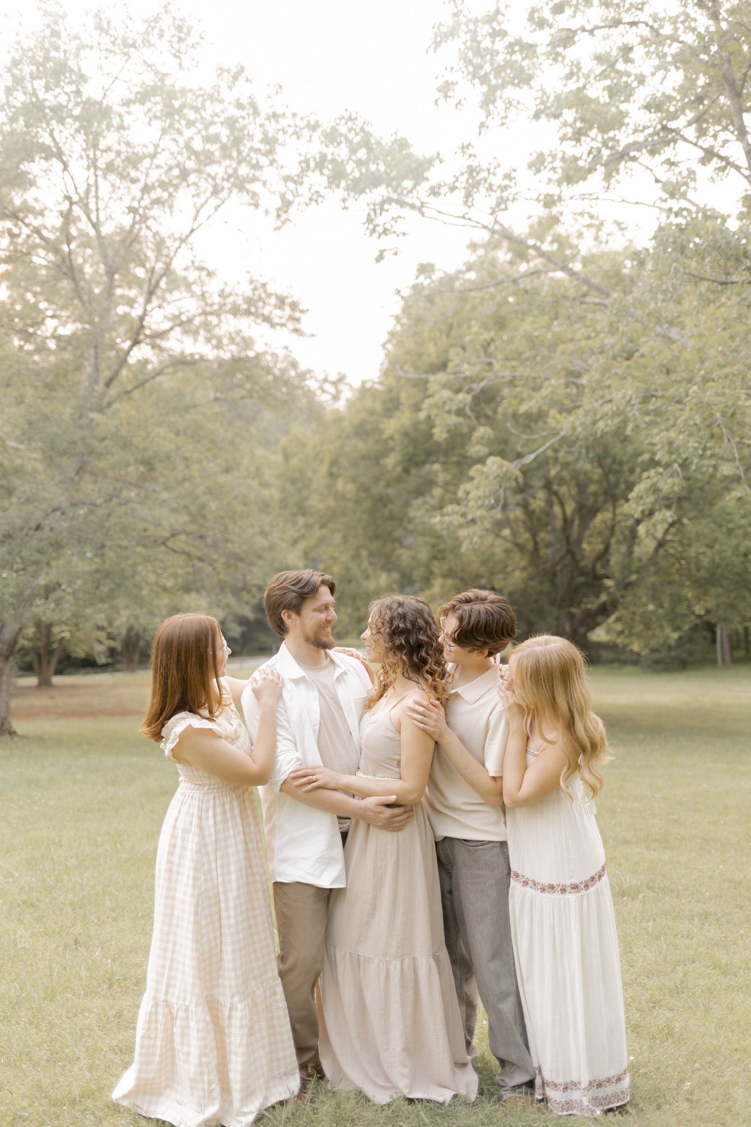 A family of five in neutral tones embracing in a lush green park during a warm outdoor photo session.