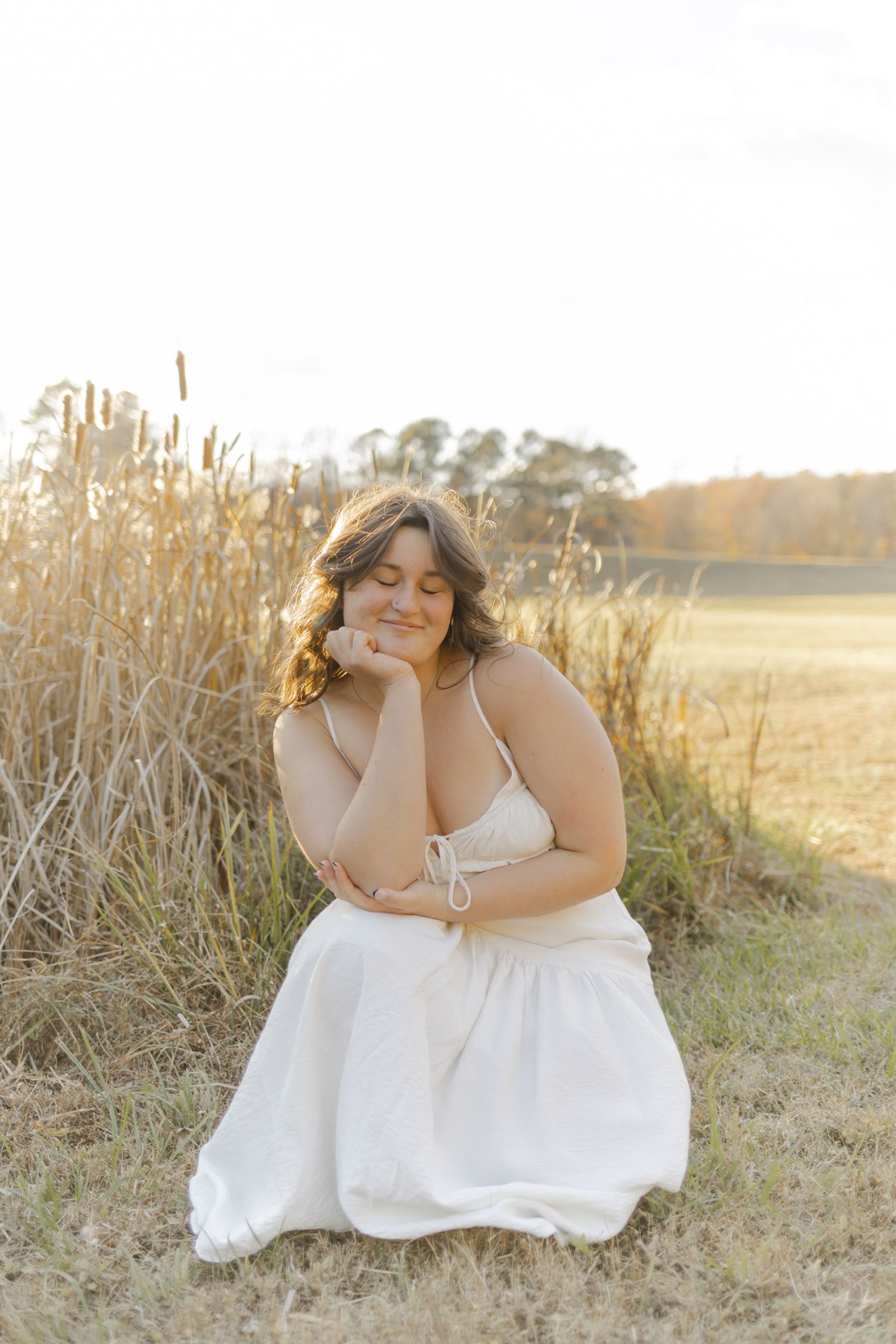 Woman in white dress kneeling peacefully in golden field at sunset, eyes closed, resting chin on hand.
