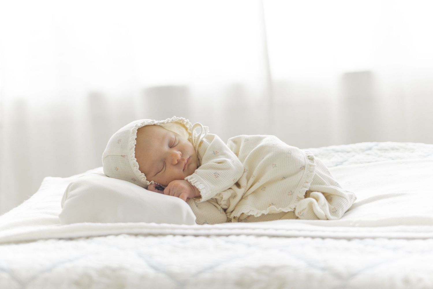 Peaceful newborn baby sleeping on white bedding, wrapped in a cream blanket and bonnet in a bright, airy room.