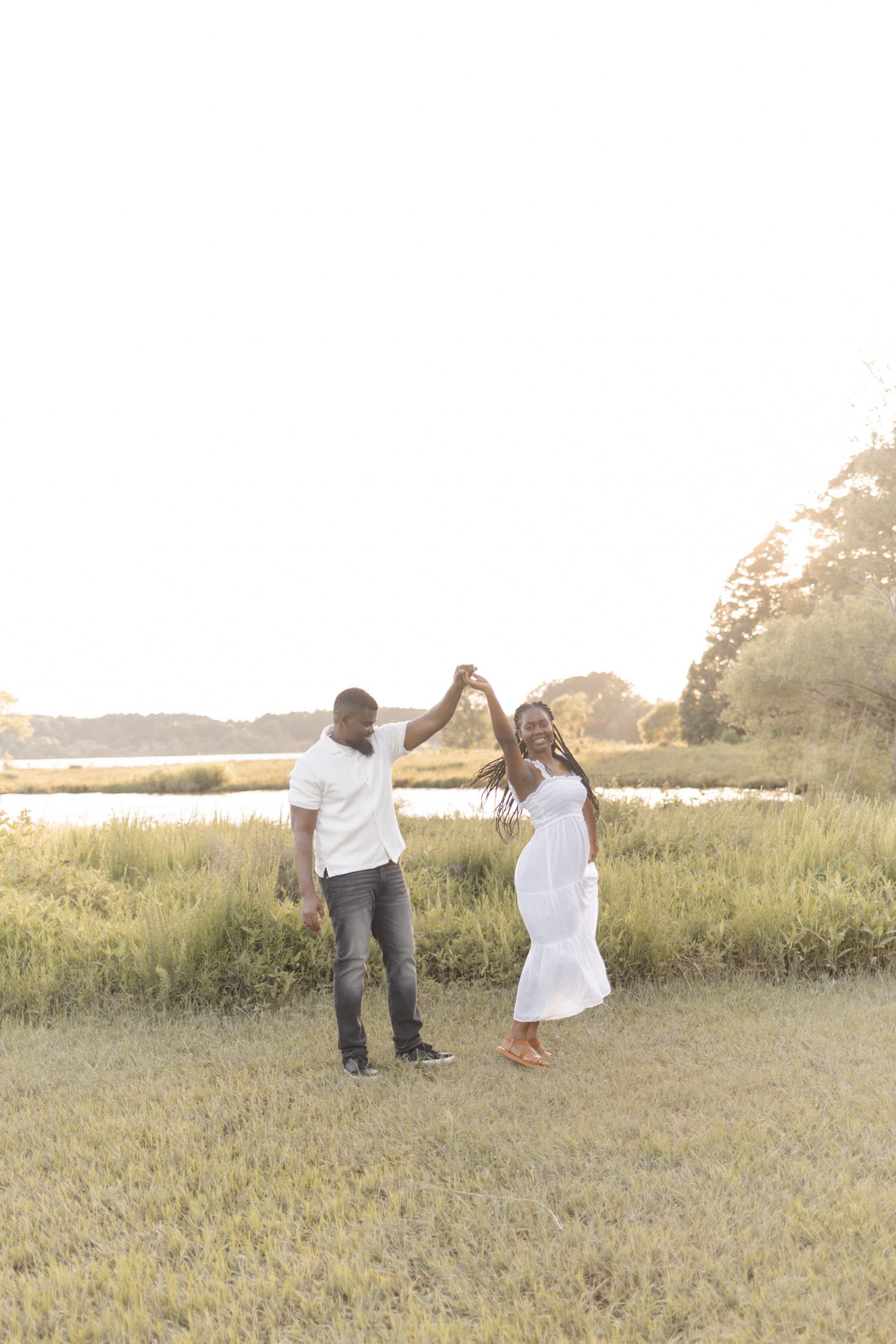 A couple dances together in a sunlit meadow near a river, the woman in a white dress and the man in a white shirt.