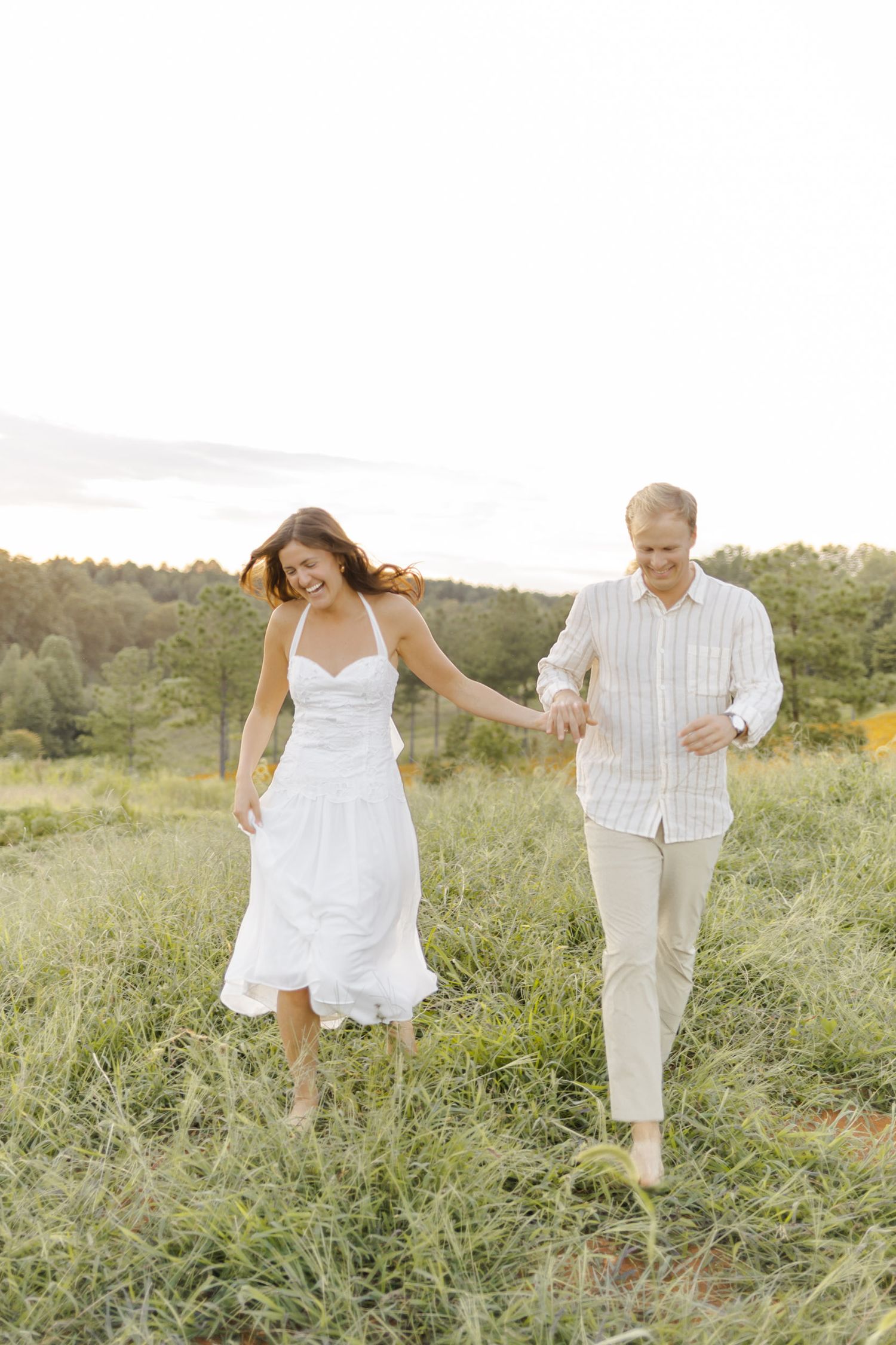 A couple in white outfits walks through a sunlit meadow holding hands at photoshoot in Chapel Hill