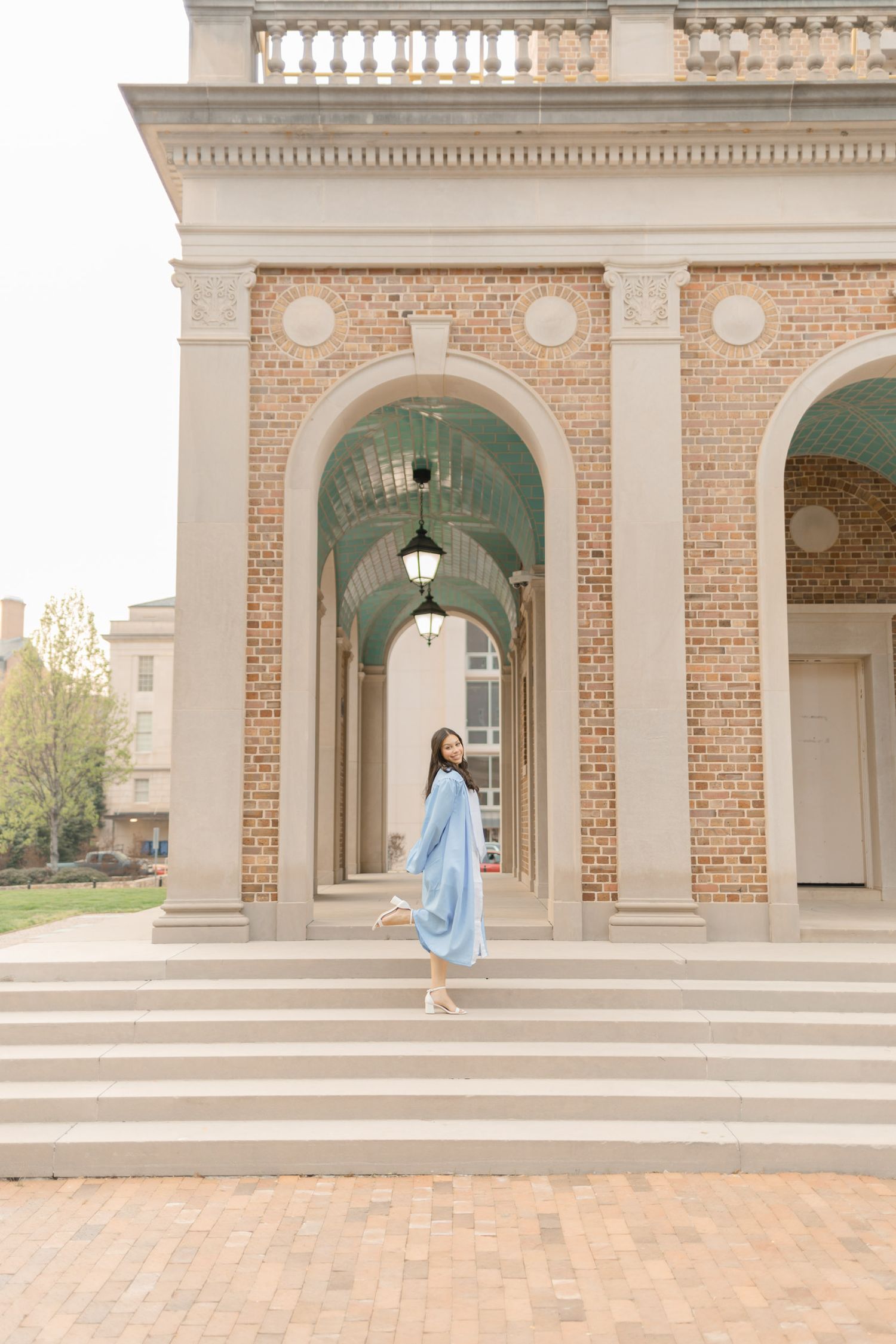 Graduate in light blue gown posing under elegant brick arches on a university campus.