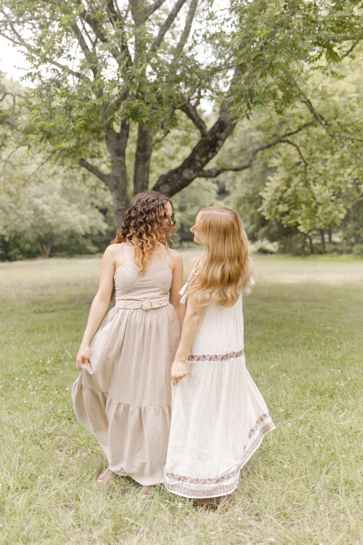 Two figures in flowing dresses walk hand in hand through tall grass in a sunlit field during photoshoot in Durham, NC.