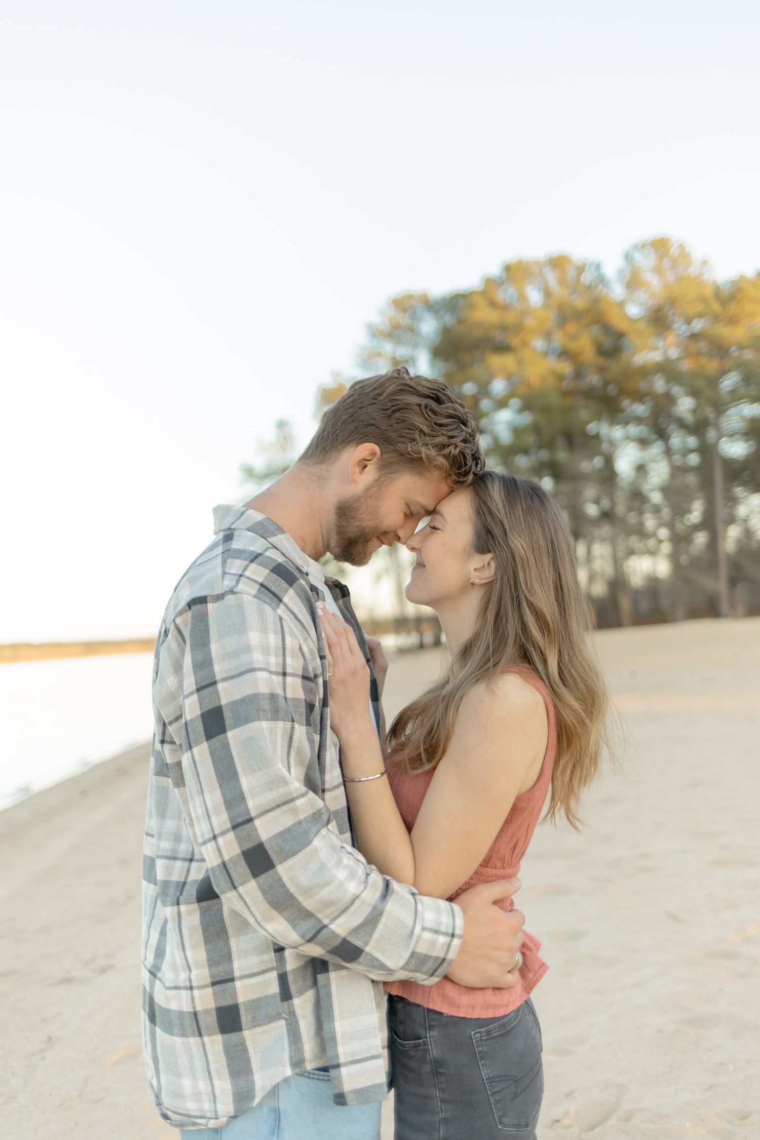 Romantic couple embraces and shares a tender moment on a sandy beach with pine trees in the background in Raleigh, North Carolina.