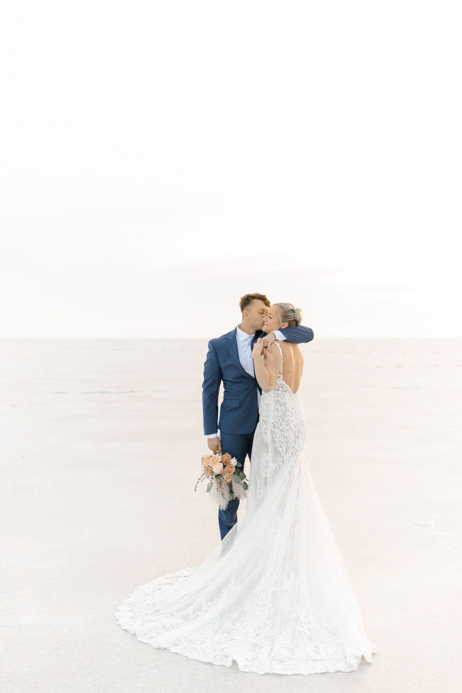 A couple in wedding attire share an intimate moment on a bright white background, with the bride wearing a lace gown.