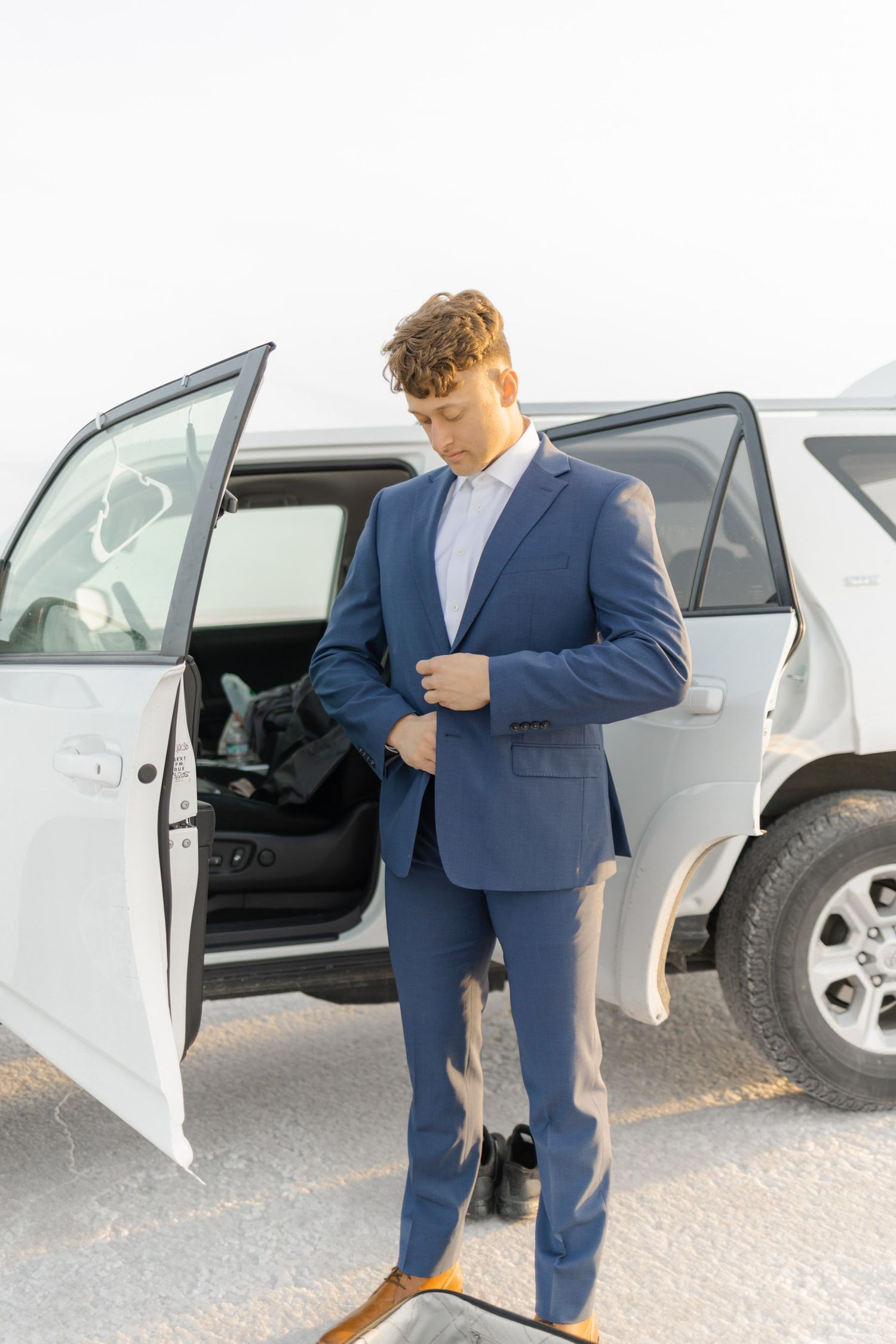 A person in a navy blue suit steps out of a white car in a parking lot.
