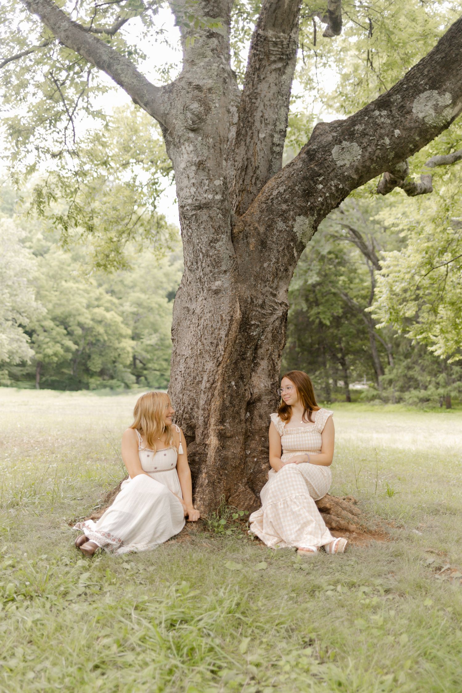 Two figures in light dresses sit beneath a large tree trunk in a serene meadow setting in Durham, North Carolina.
