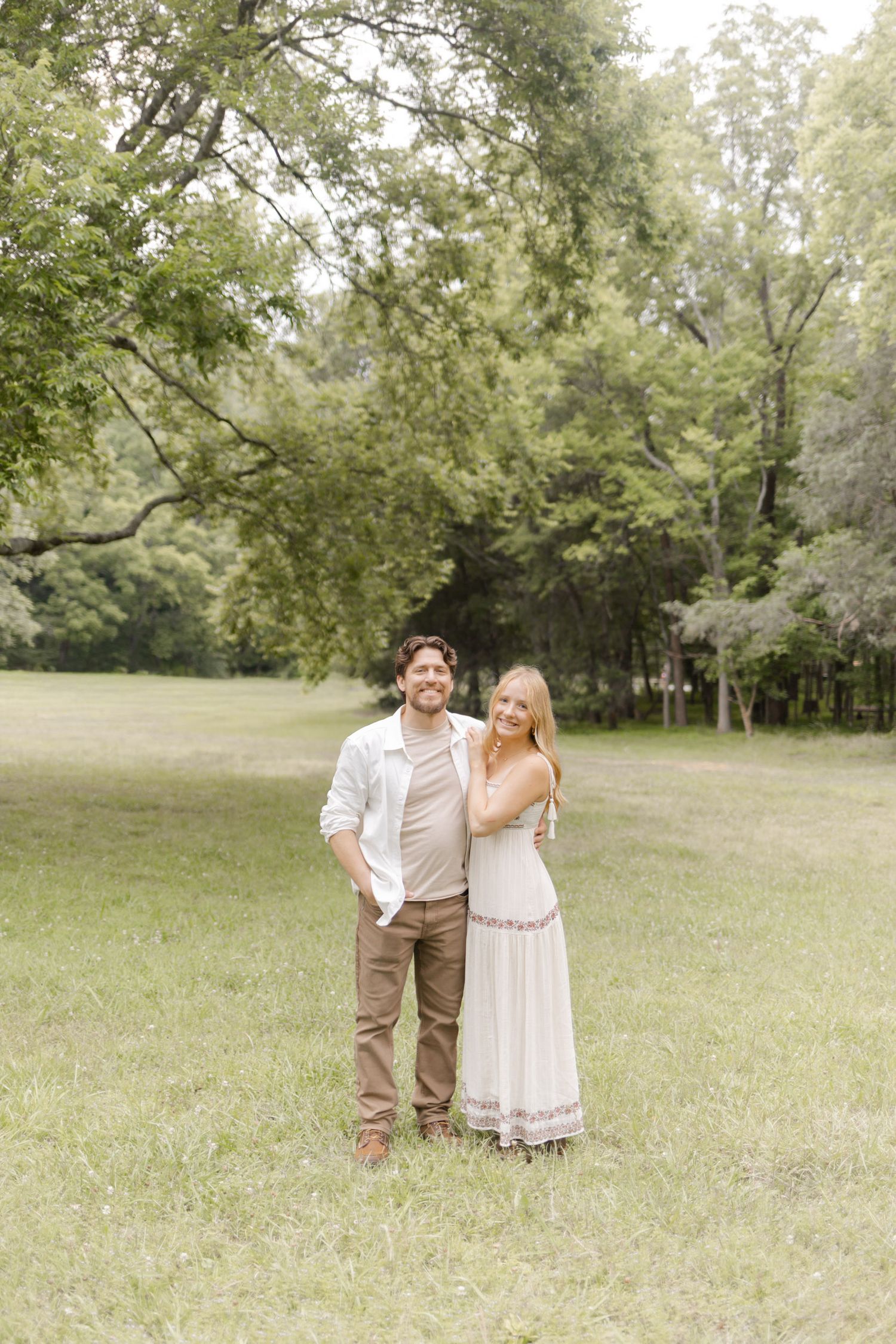A couple poses together in a sunlit meadow wearing coordinated neutral tones and white dresses during photoshoot in Durham, NC.