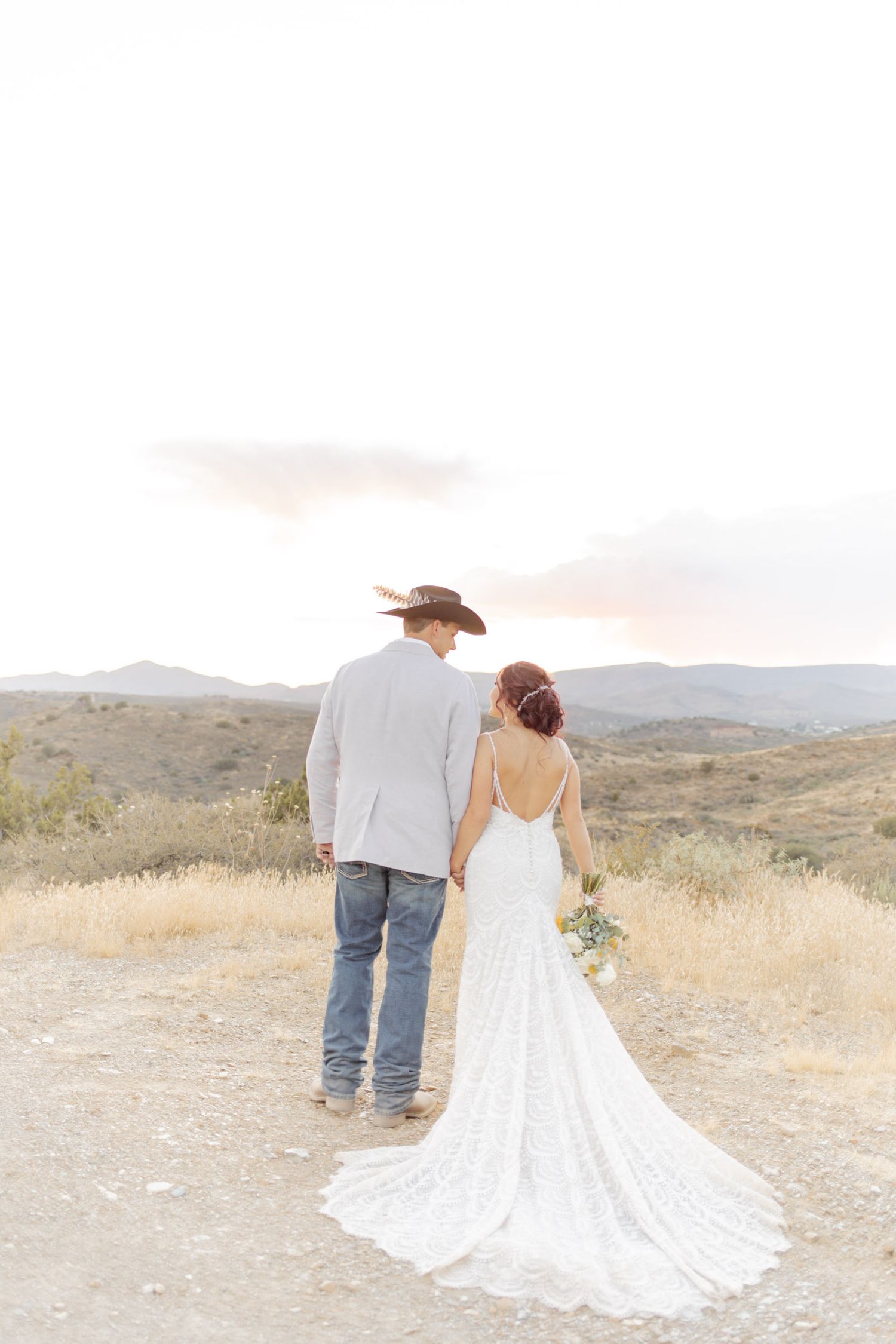 A bride and groom hold hands facing a scenic mountain landscape, she in a lace gown, he in jeans and cowboy hat.