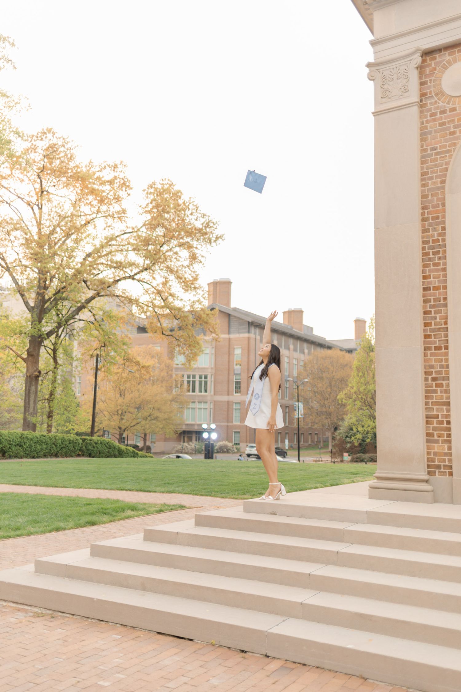 A graduate in a white dress tosses her cap in the air on university steps surrounded by autumn trees.