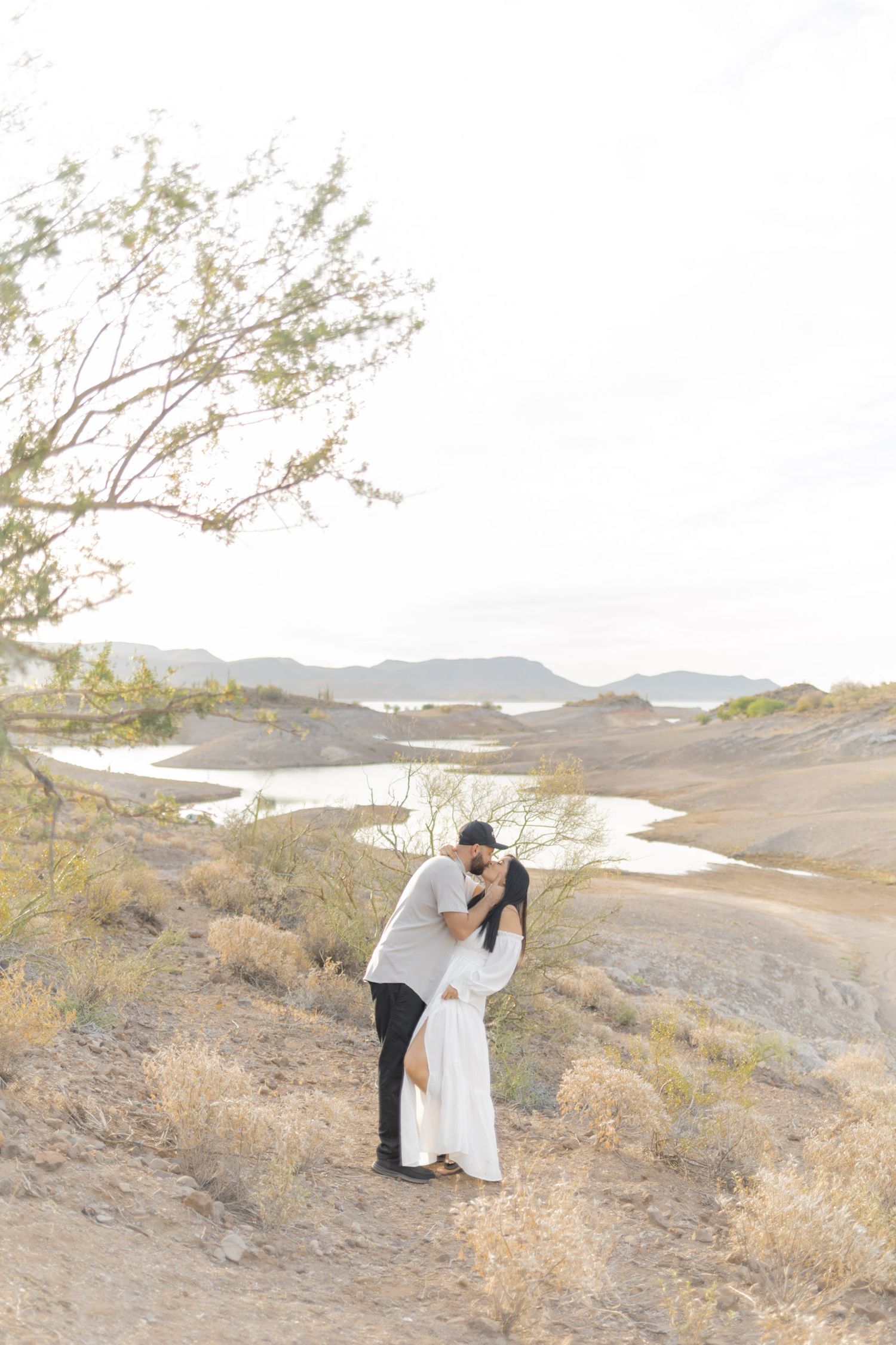 Couple sharing a romantic kiss on arid desert hillside with scenic lake and mountain landscape in background.