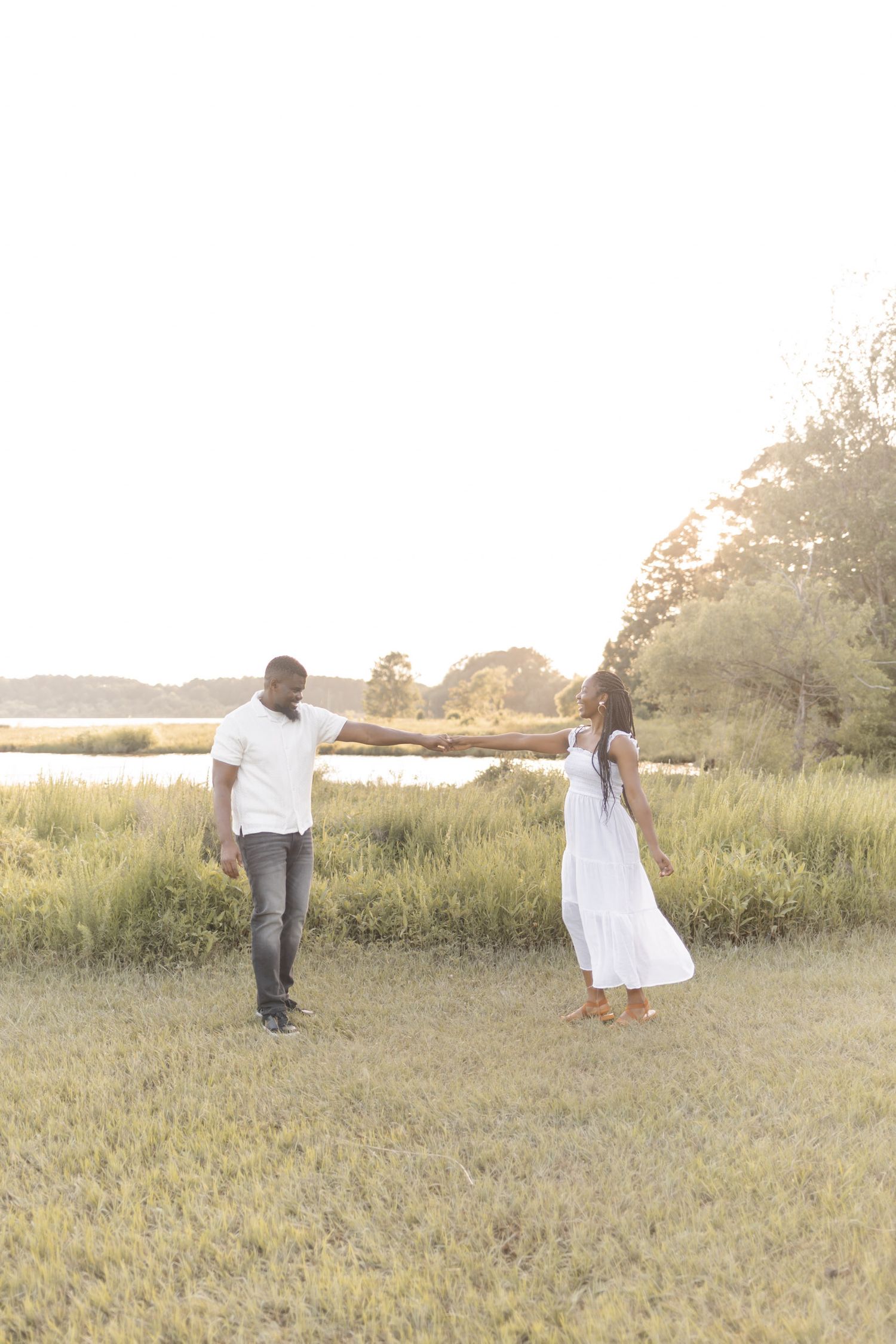 A couple in white outfits holds hands while dancing in a sunlit grassy field near a serene waterway.