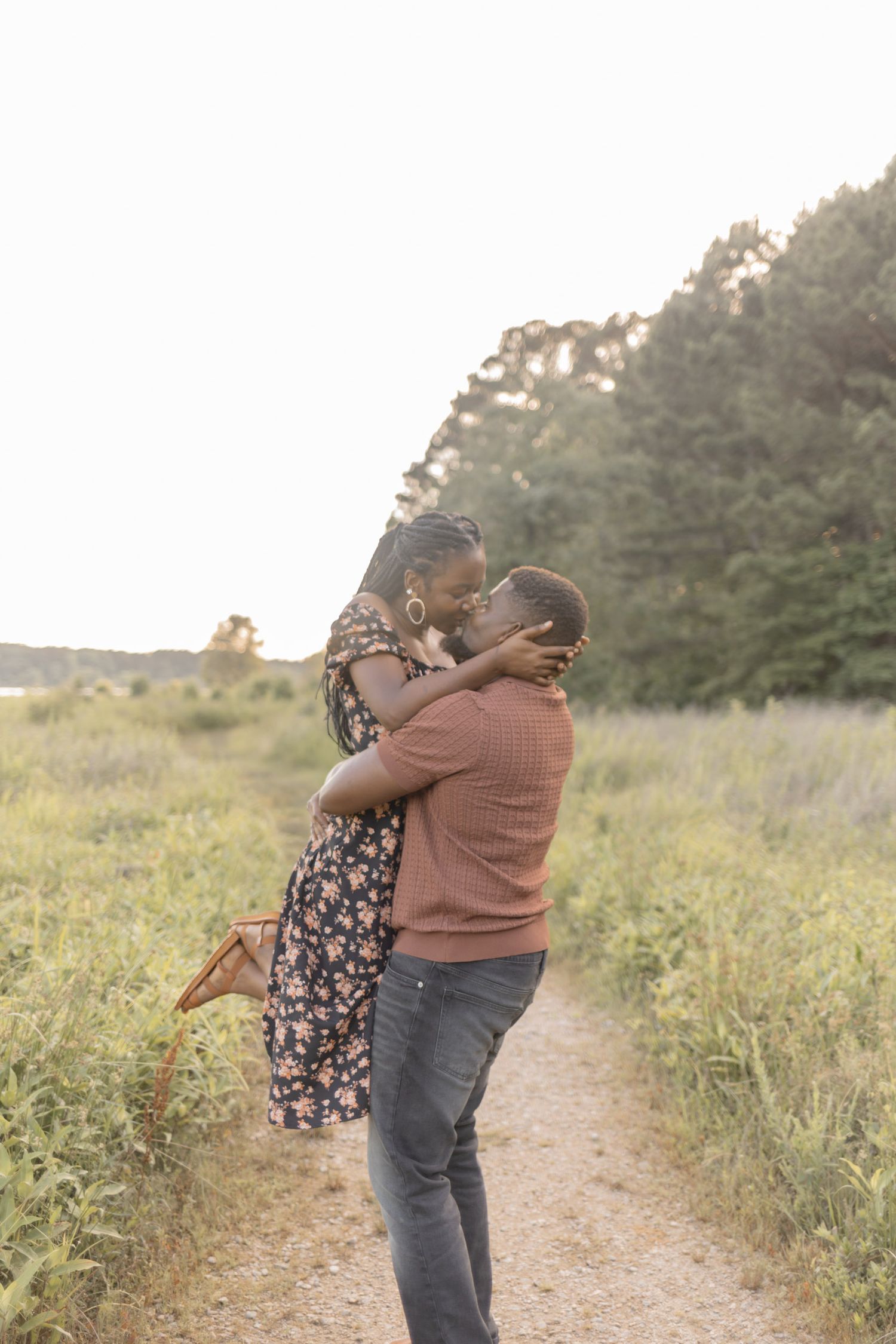 Man lifting woman in floral dress as they embrace on a nature trail surrounded by lush green fields.