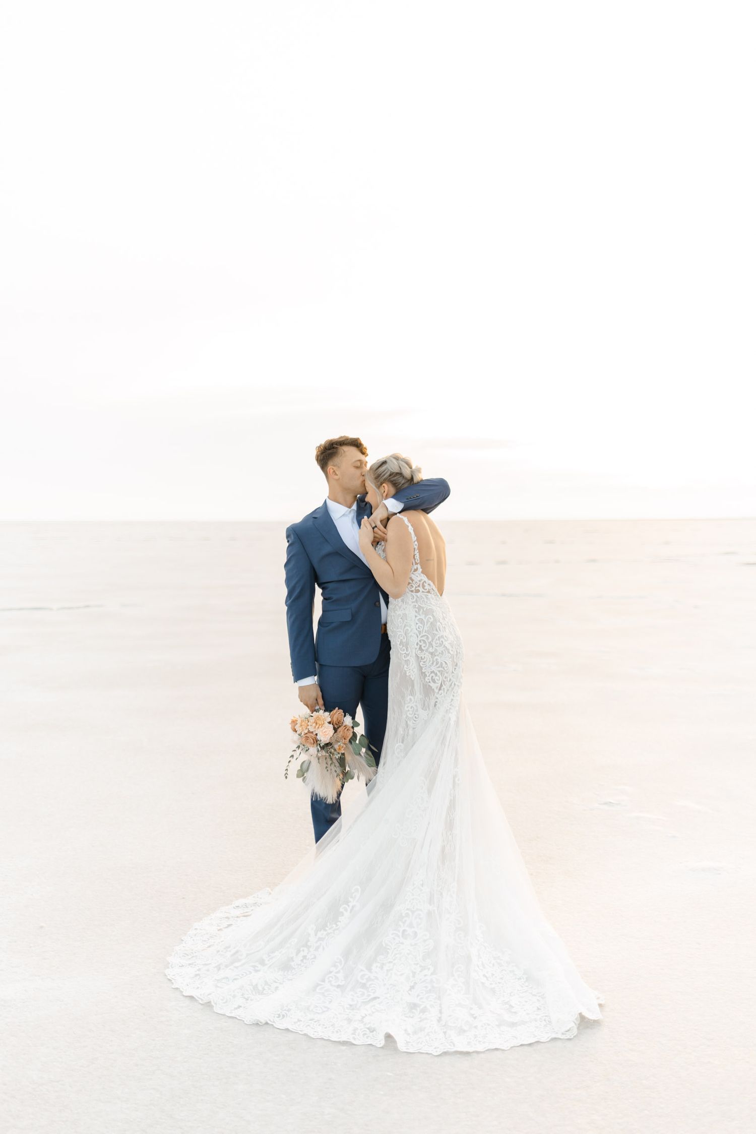 A couple in wedding attire share an intimate dance pose on a bright desert landscape.
