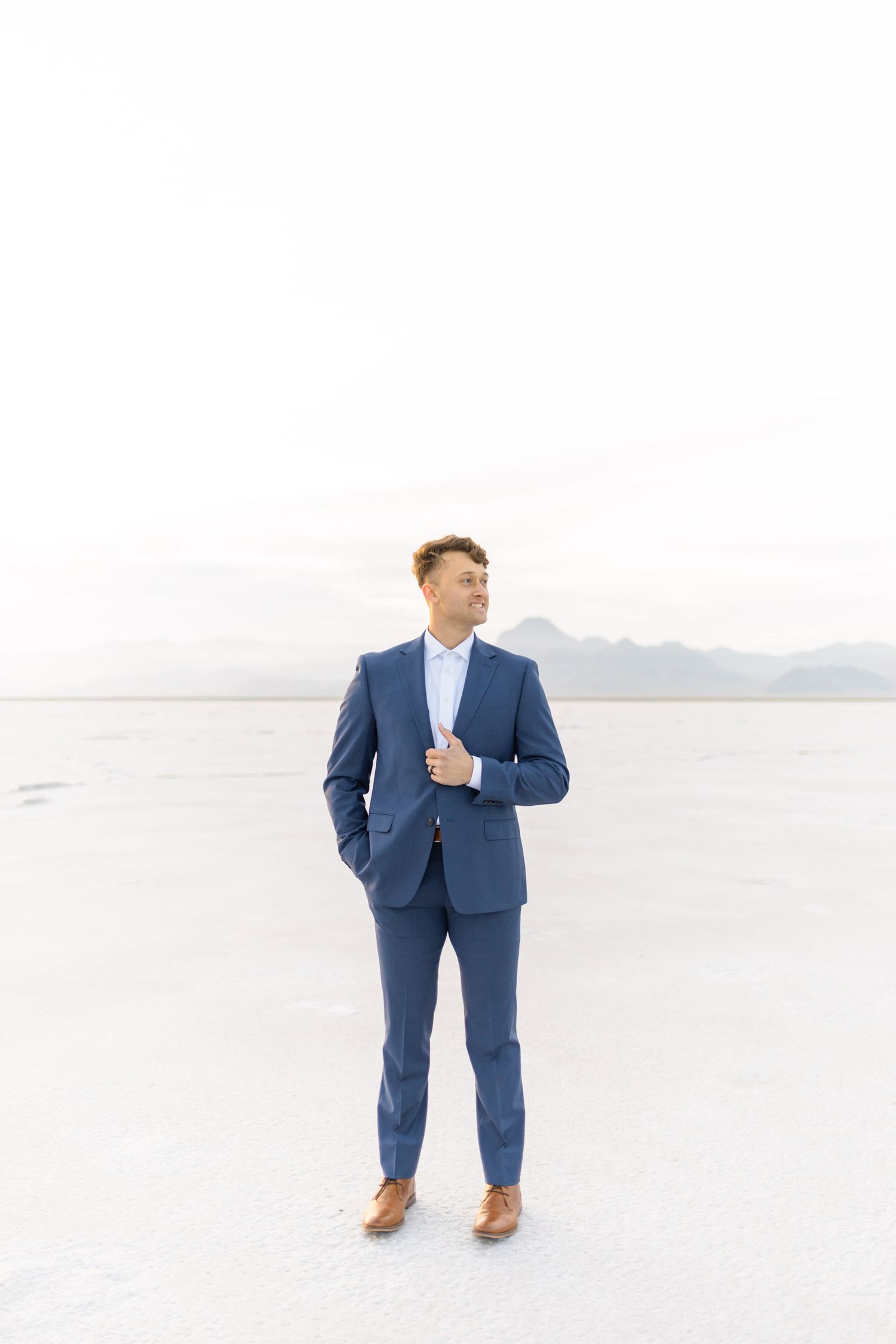 Person in navy blue suit stands alone on vast white salt flats with mountains on the horizon.