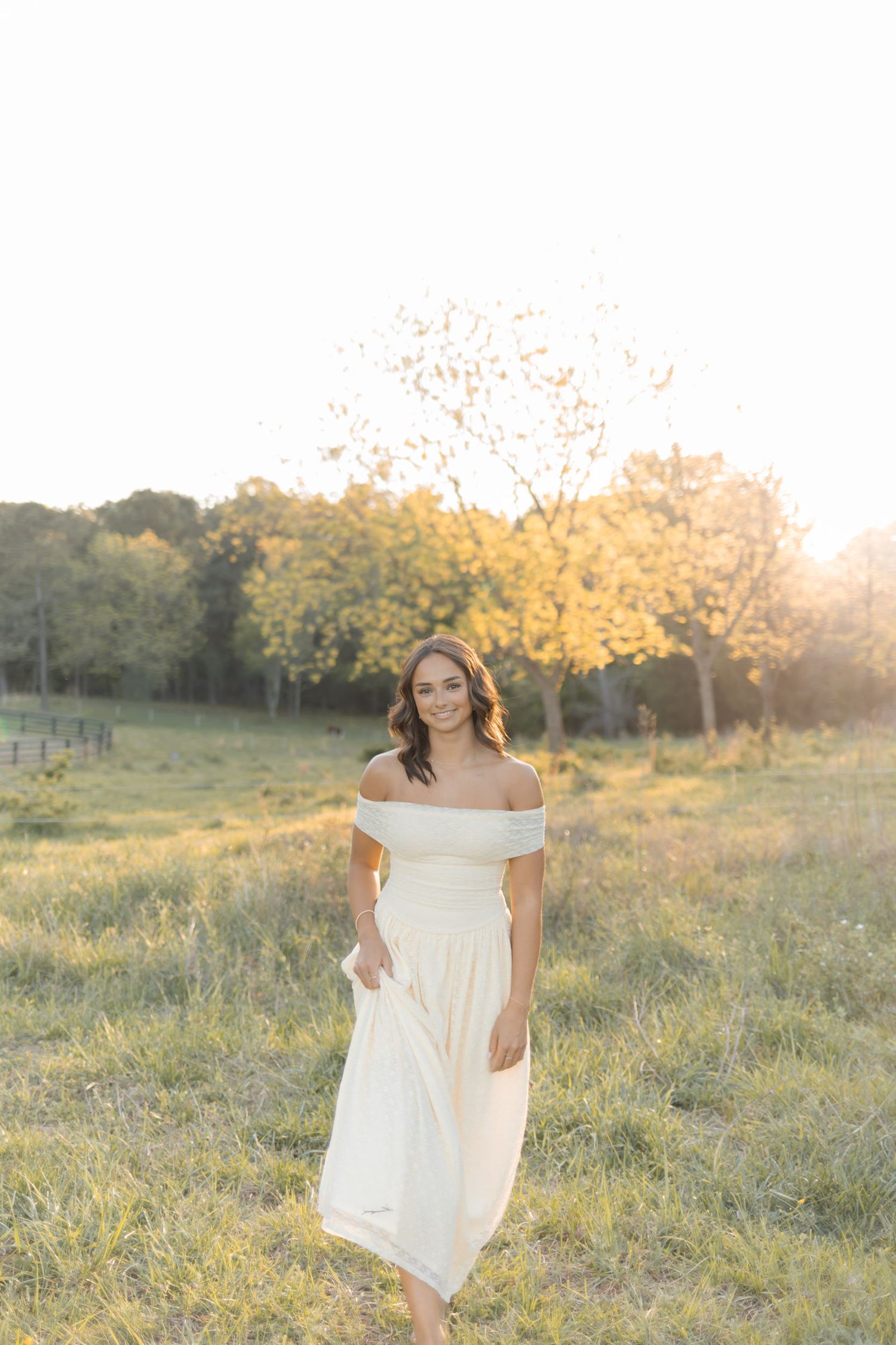 Woman in white off-shoulder midi dress walking through golden sunlit meadow.