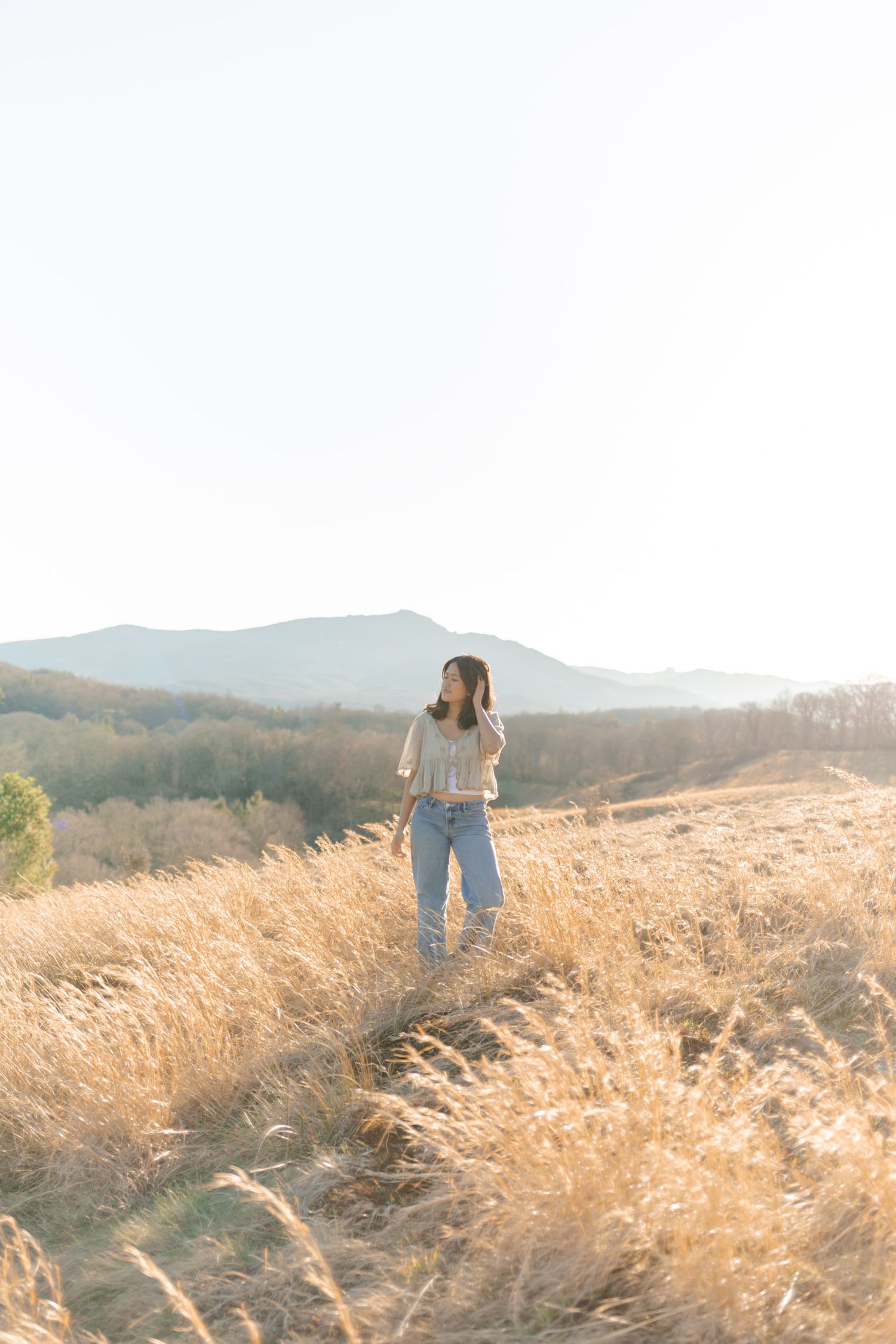 Young woman standing in tall golden grass field with misty mountain backdrop during golden hour.