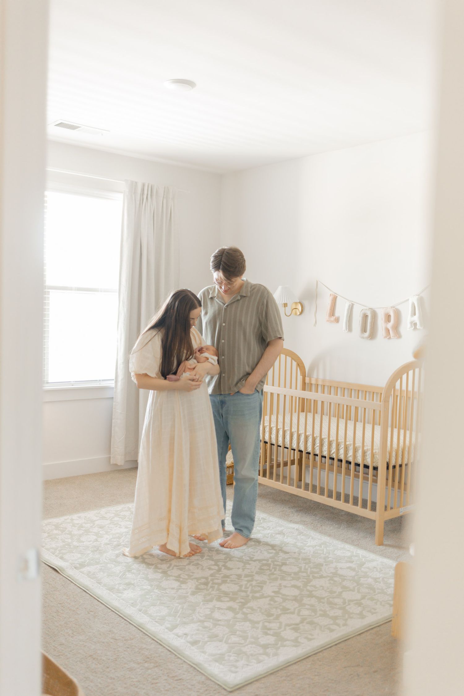 New parents stand together in a softly lit nursery holding their newborn near a wooden crib with a name banner spelling Lora.