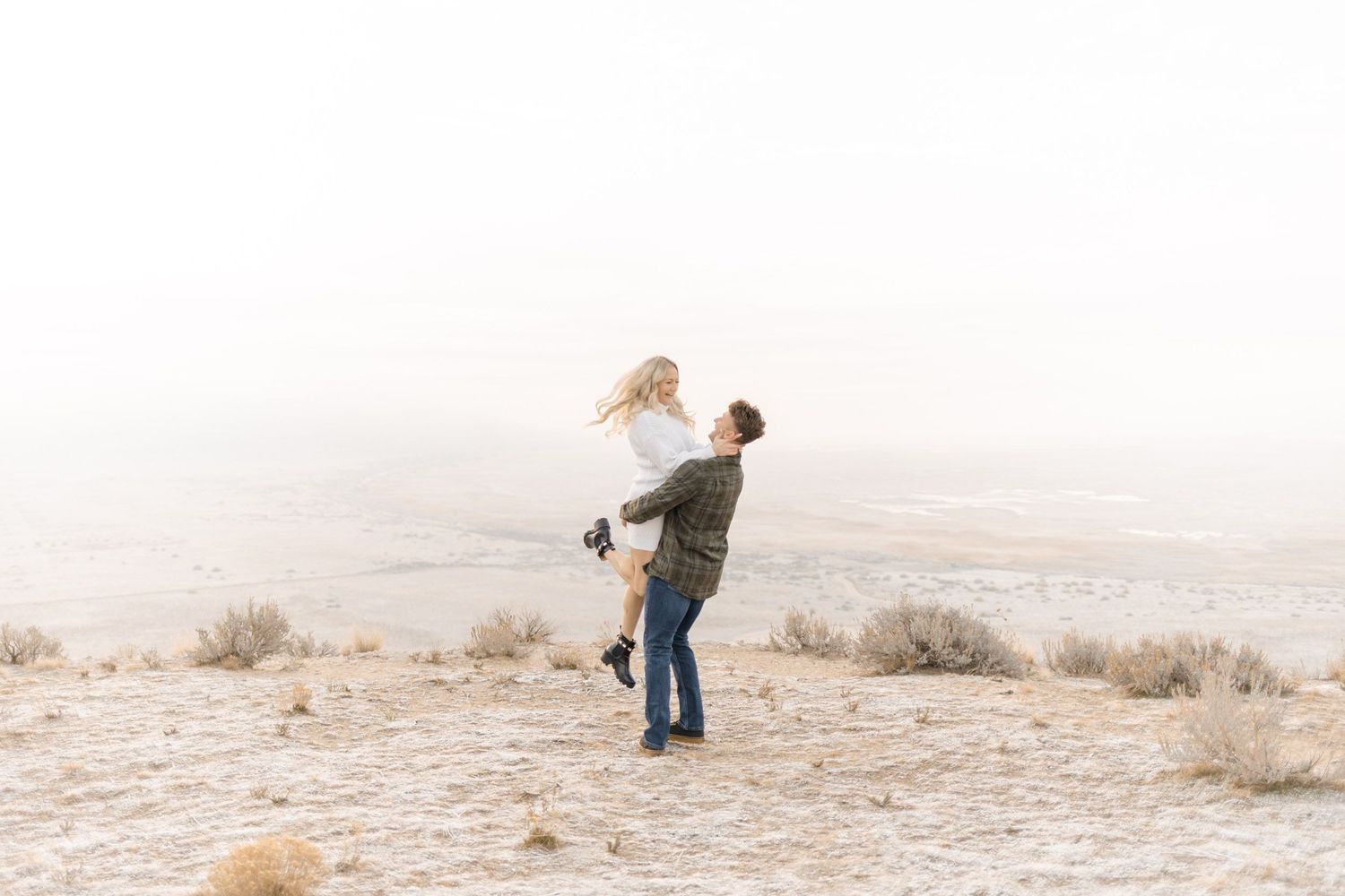 A man joyfully lifts a woman in a snowy desert landscape, capturing a romantic couples engagement photo.