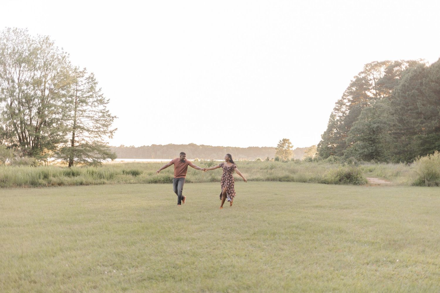 Couple holding hands and running through a lush green field surrounded by trees at golden hour.