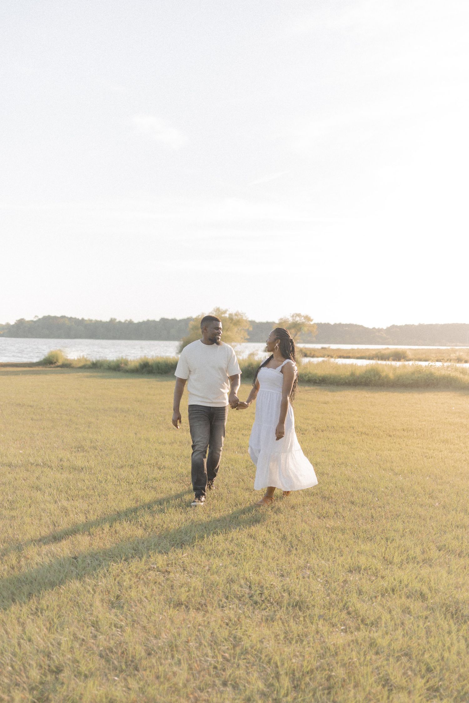 A couple holds hands walking through a sunlit green field near a waterfront, woman in white dress, man in white shirt.