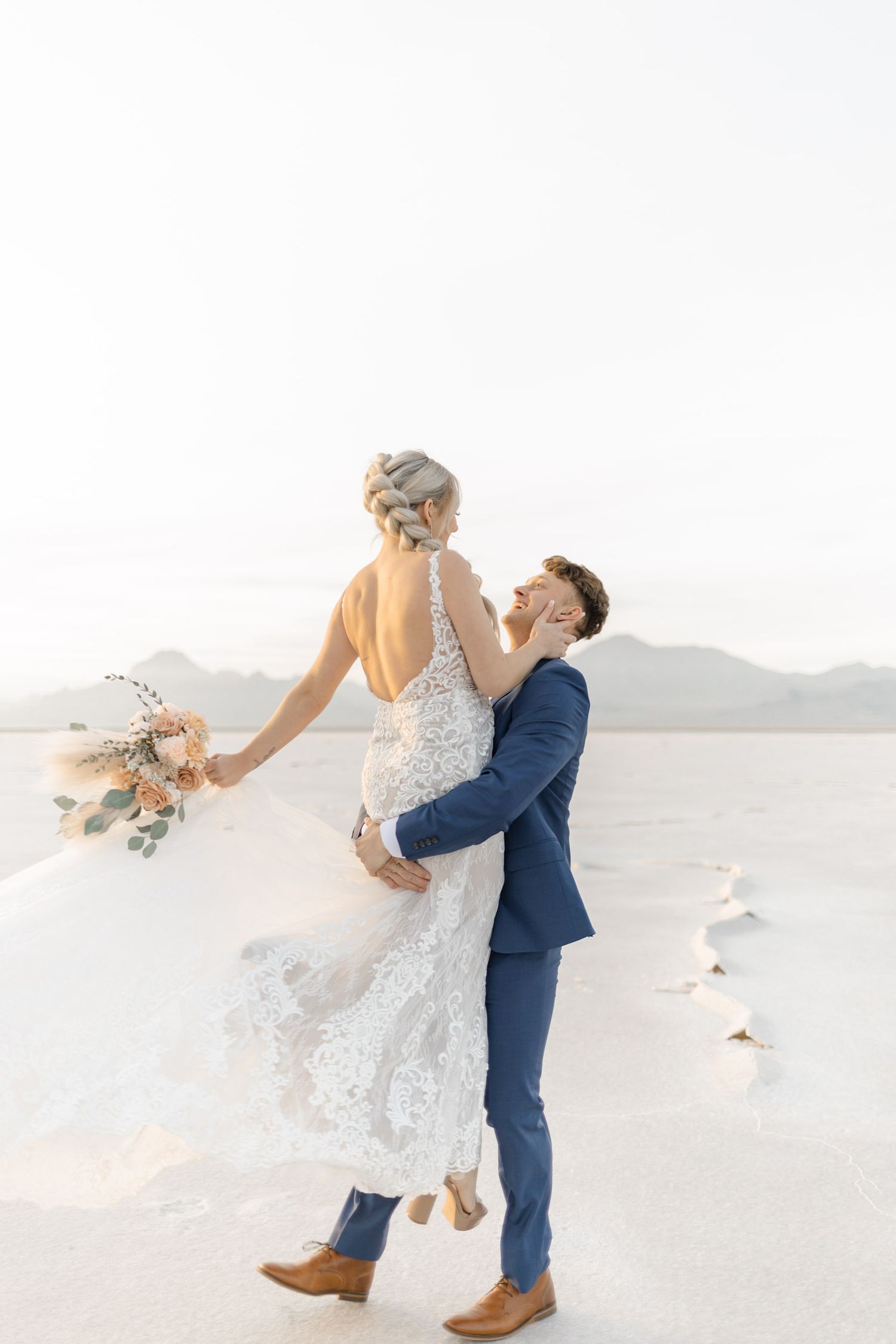 Newlyweds embrace and spin together on a bright desert landscape during their wedding photoshoot.