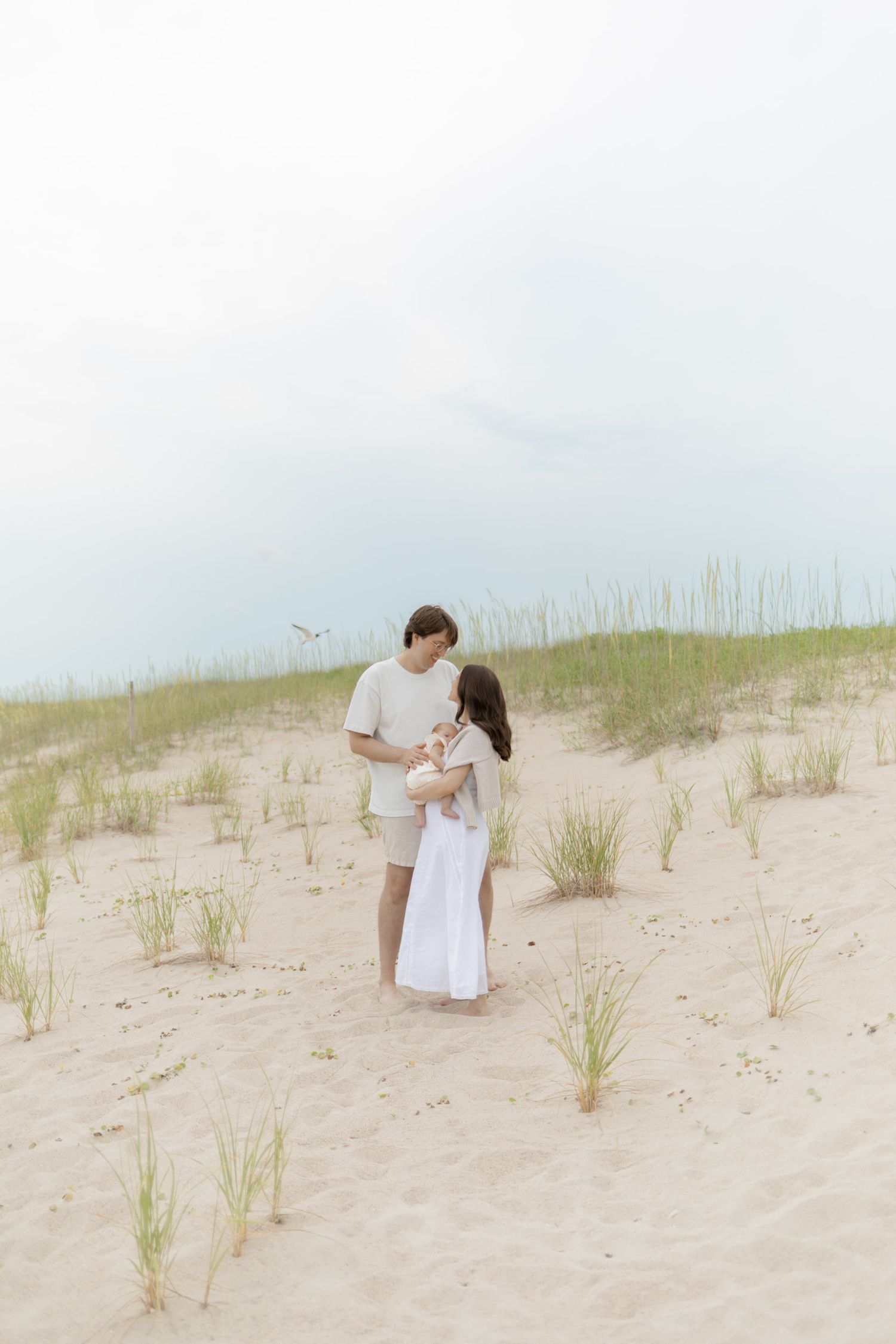 A couple stands together on sandy beach dunes with sea grass, dressed in neutral tones, sharing a tender moment.