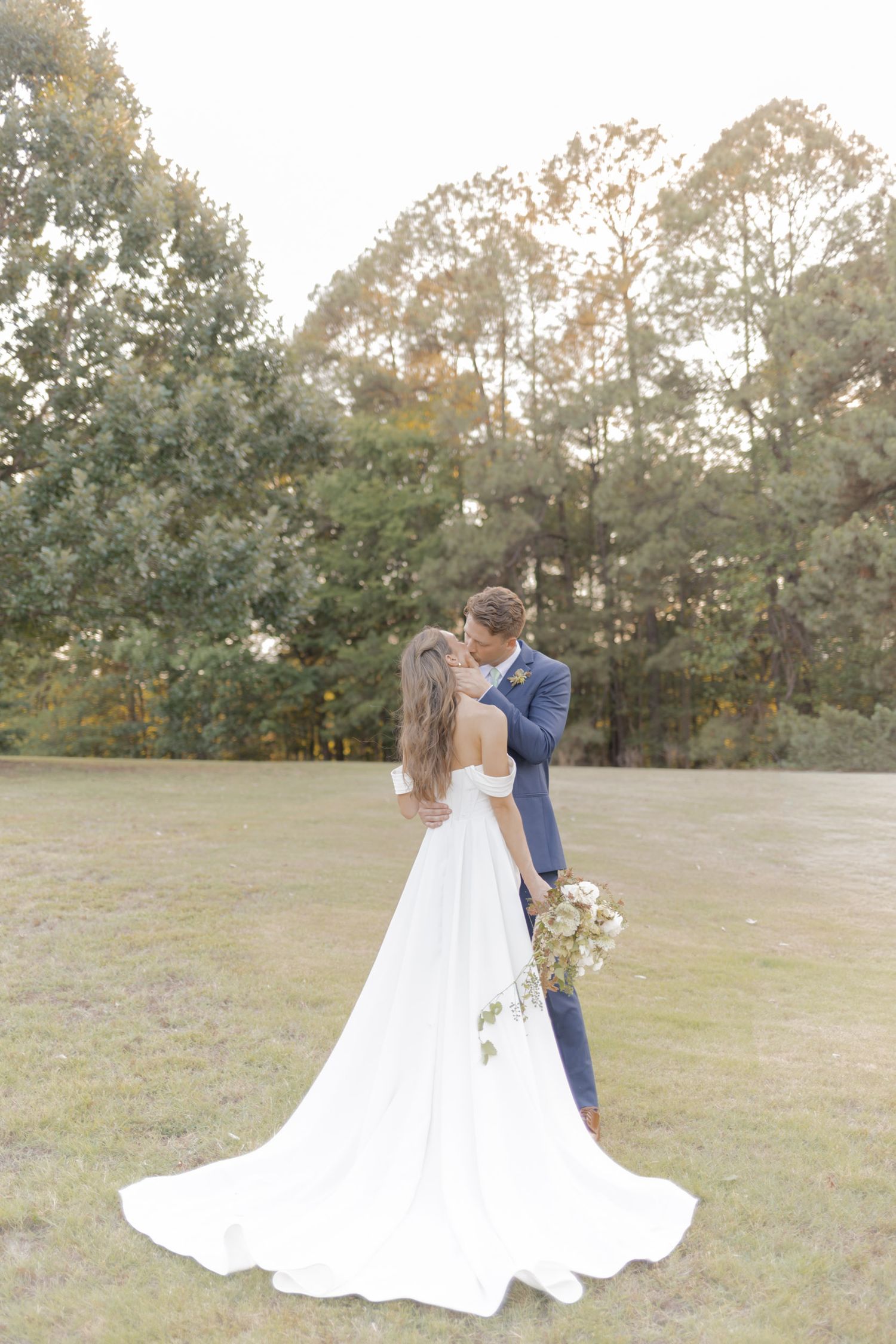 Bride and groom sharing a kiss in an open field, bride wearing an elegant off-shoulder white gown with a long train.