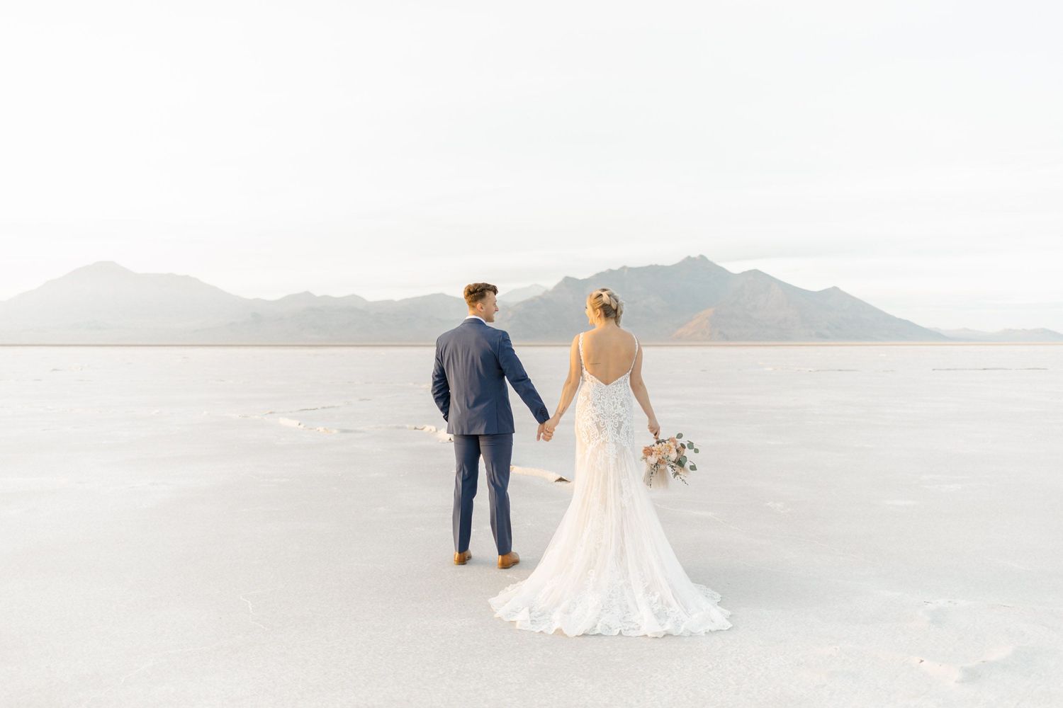Elegant wedding couple walking along a serene beach with mountains in the distance.