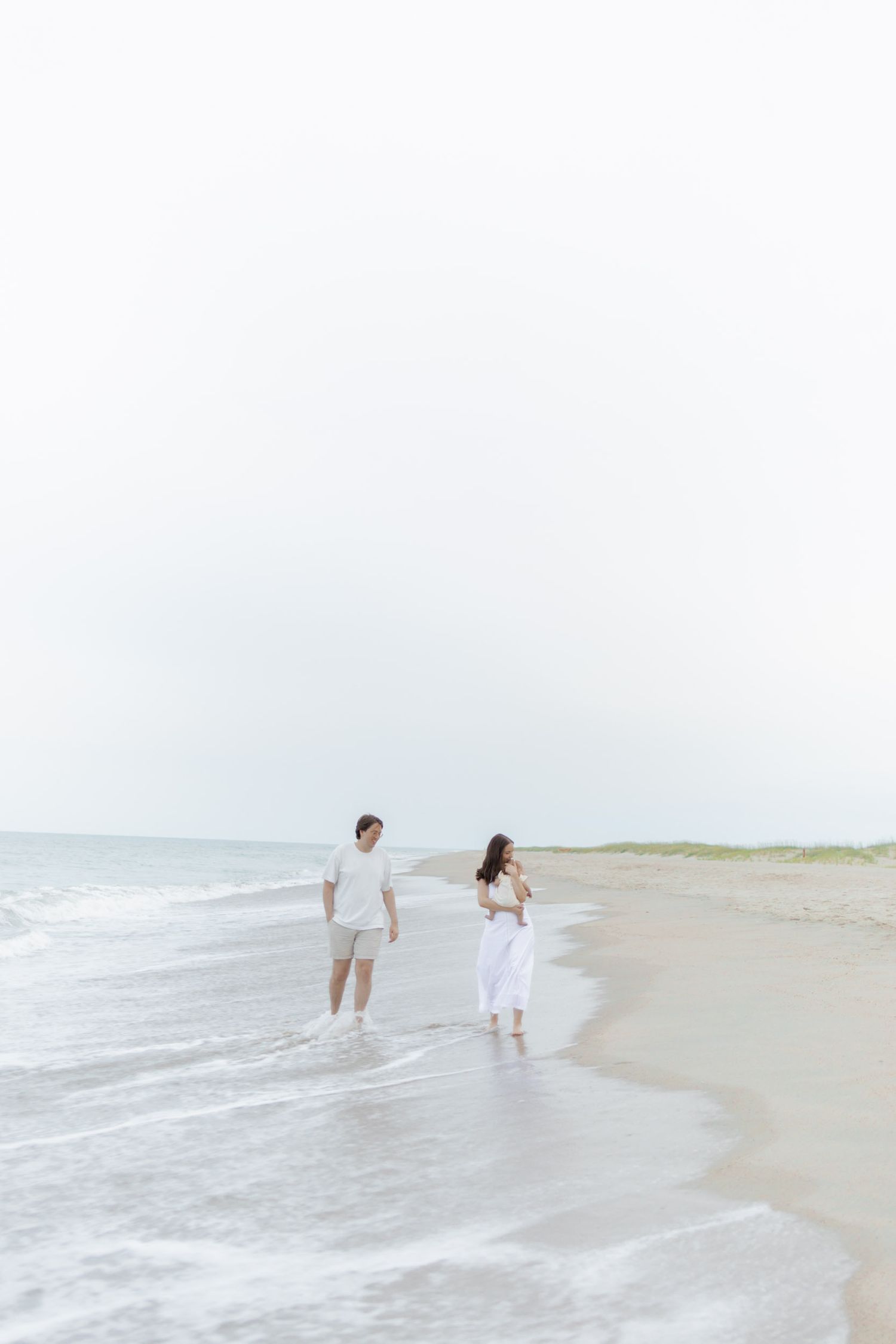 A couple walks along the shoreline at the beach, dressed in white and light gray outfits, with gentle waves at their feet.