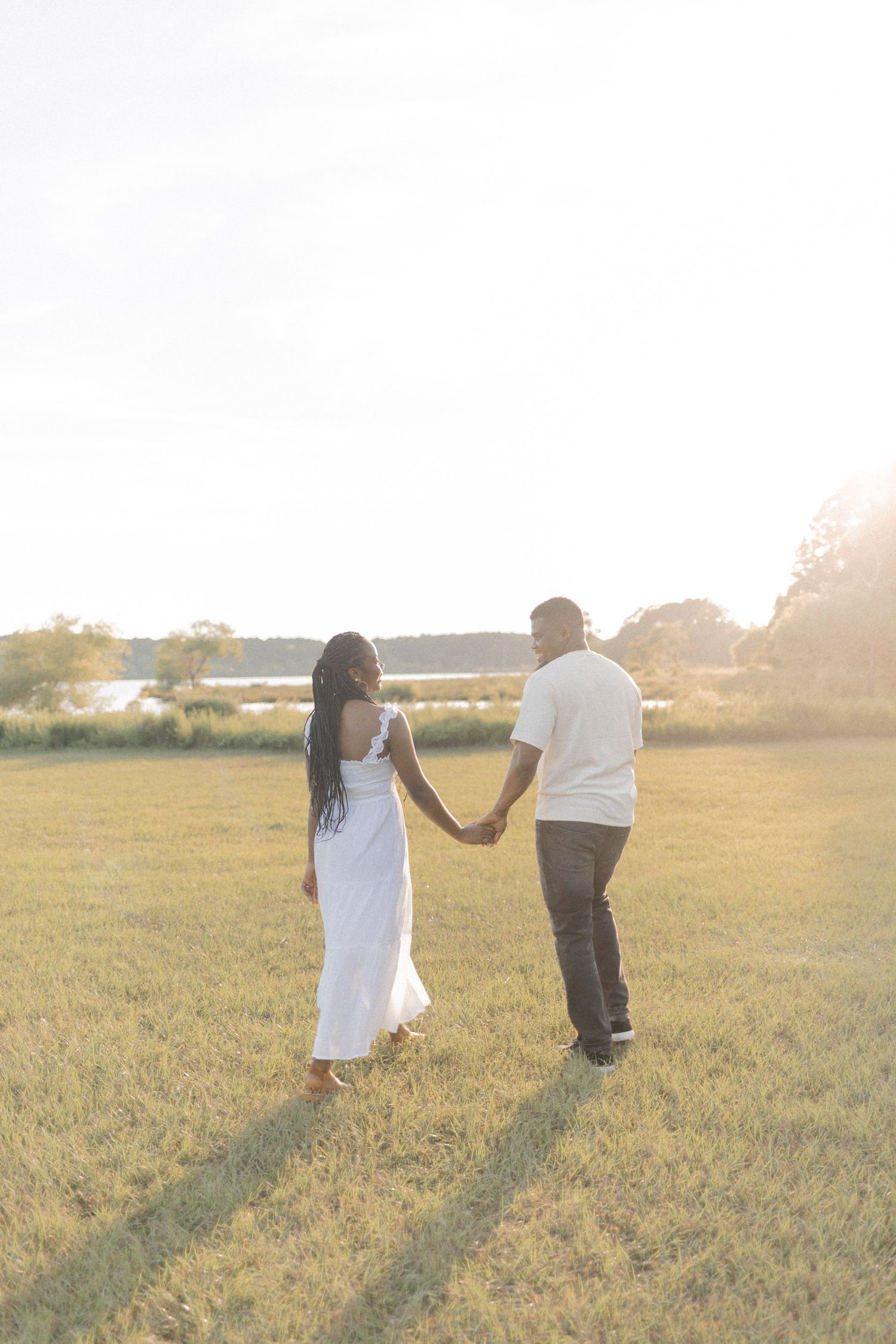 Couple holding hands walking through a sunlit green field near a lake, woman in white dress, man in white shirt.
