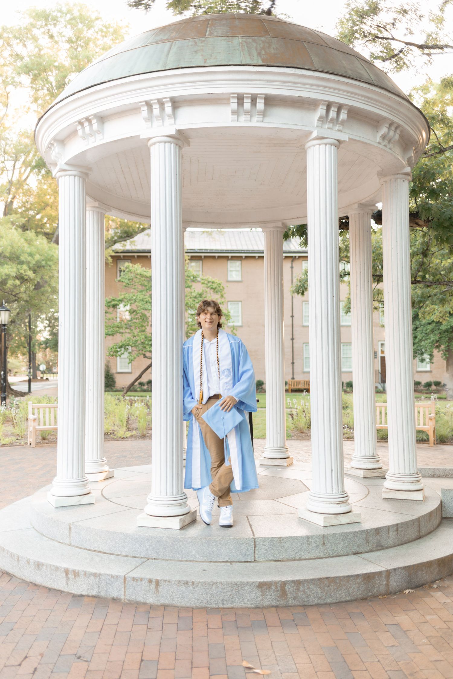Graduate in light blue cap and gown posing at the Old Well rotunda at UNC Chapel Hill.