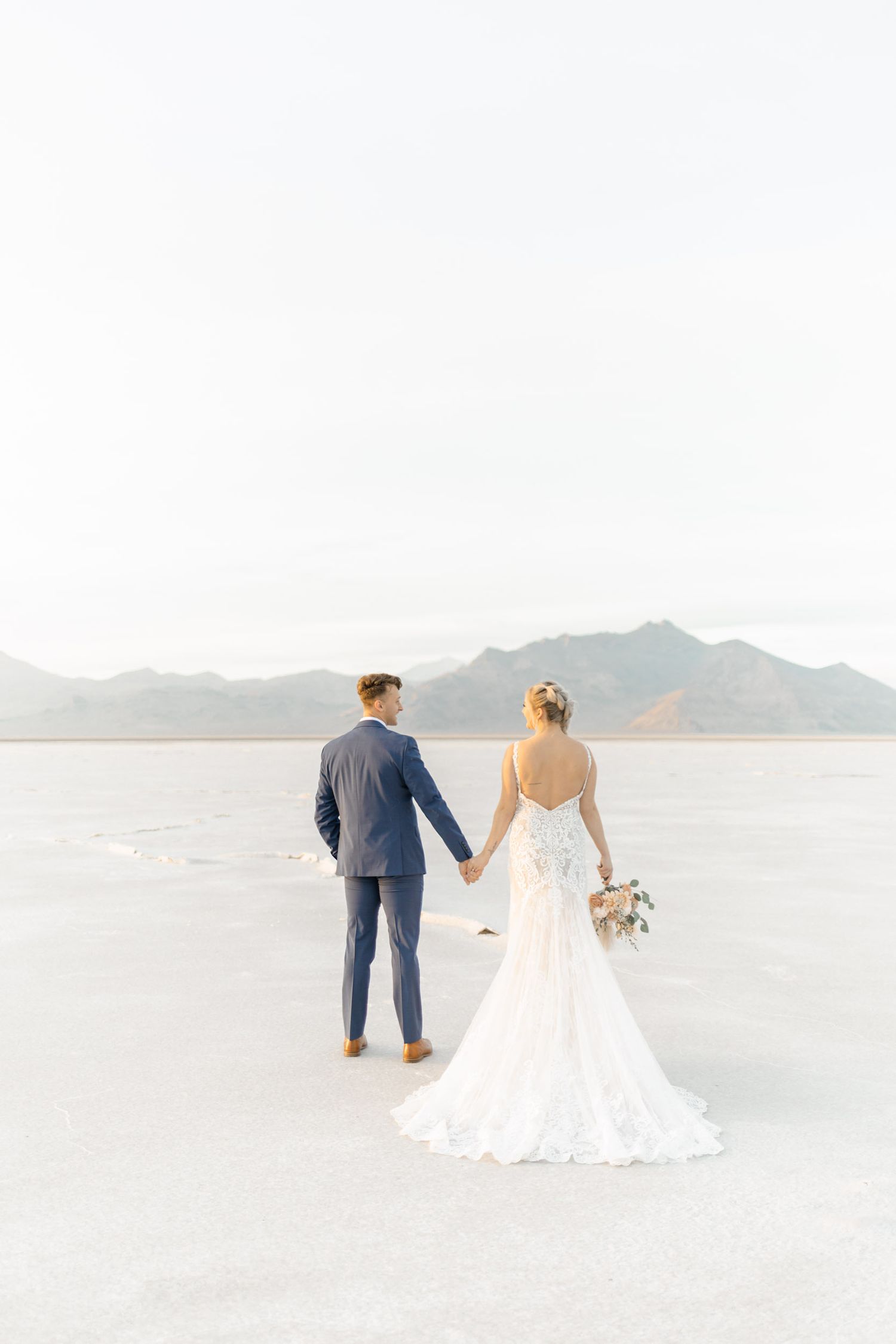 A couple in wedding attire walks hand in hand along a serene beach with mountains in the distance.