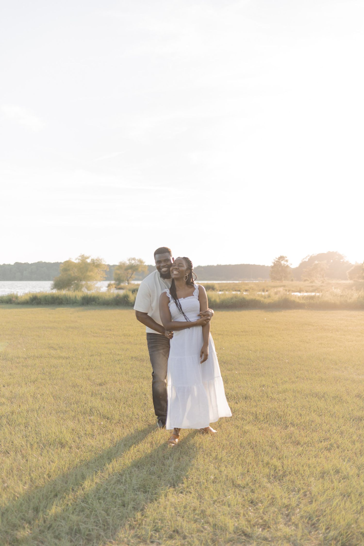 A couple embraces in a sunlit green field near a lake, woman in white dress, golden hour glow.