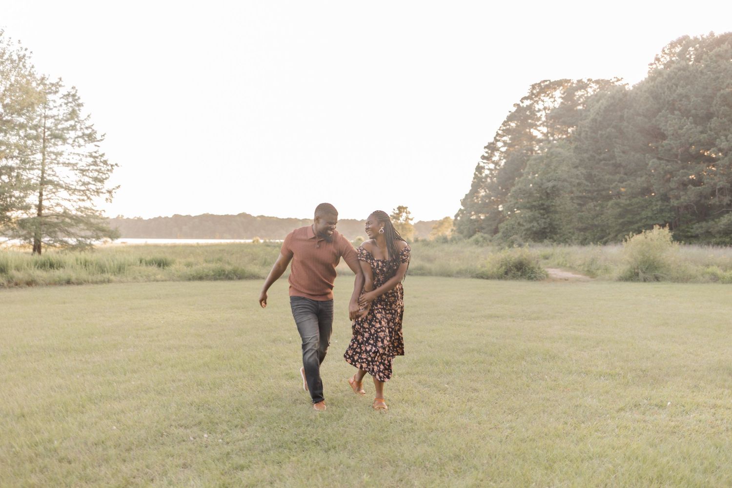 A couple joyfully runs through a sunlit green field surrounded by trees during a romantic outdoor engagement session.
